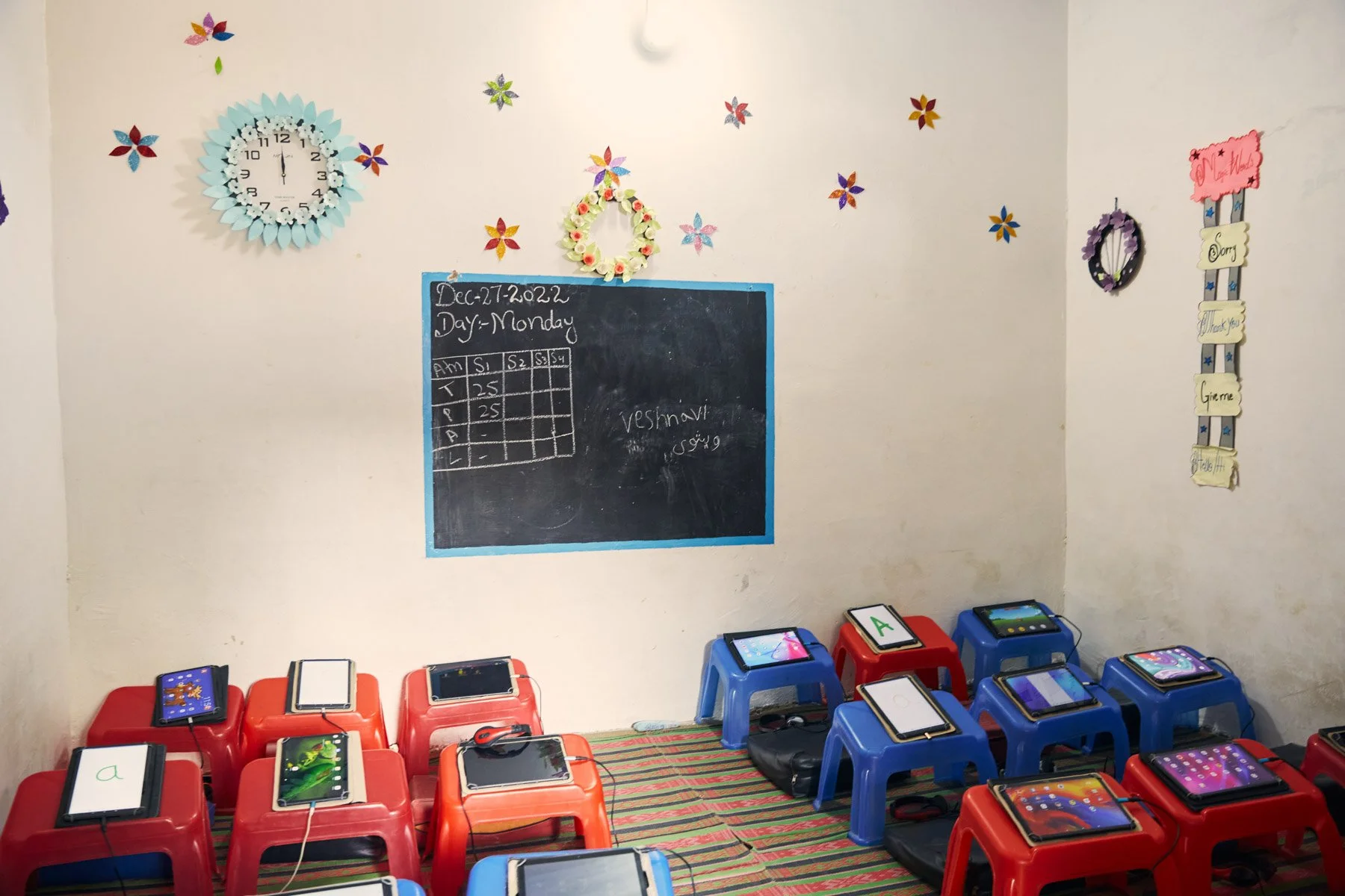Classroom with small blue and red stools, each holding a tablet, and decorated walls with a clock, floral decorations, and a chalkboard with writing and a calendar. For Teach the World Foundation, Razzaqabad, Karachi, Pakistan.