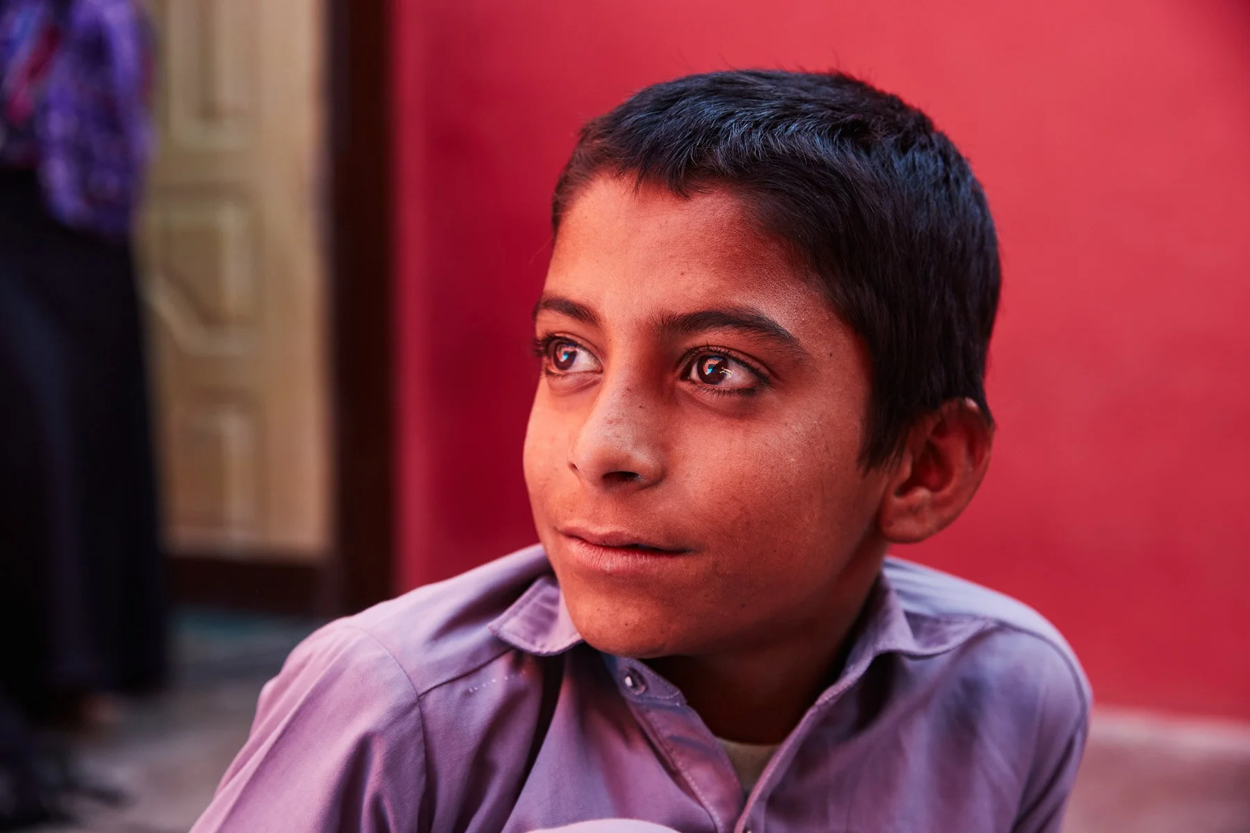 Close-up of a young boy with short dark hair, light brown skin, and blue eyes, looking to the left with a thoughtful expression. For Teach the World Foundation, Razzaqabad, Karachi, Pakistan.