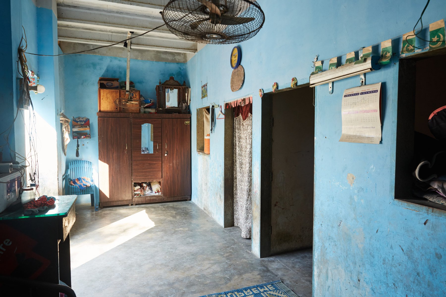 Interior of a room with blue walls, a wooden wardrobe, a small blue plastic chair, a blue and red patterned rug on the concrete floor, and a ceiling fan. Sunlight coming through a window. For Teach the World Foundation, Razzaqabad, Karachi, Pakistan.
