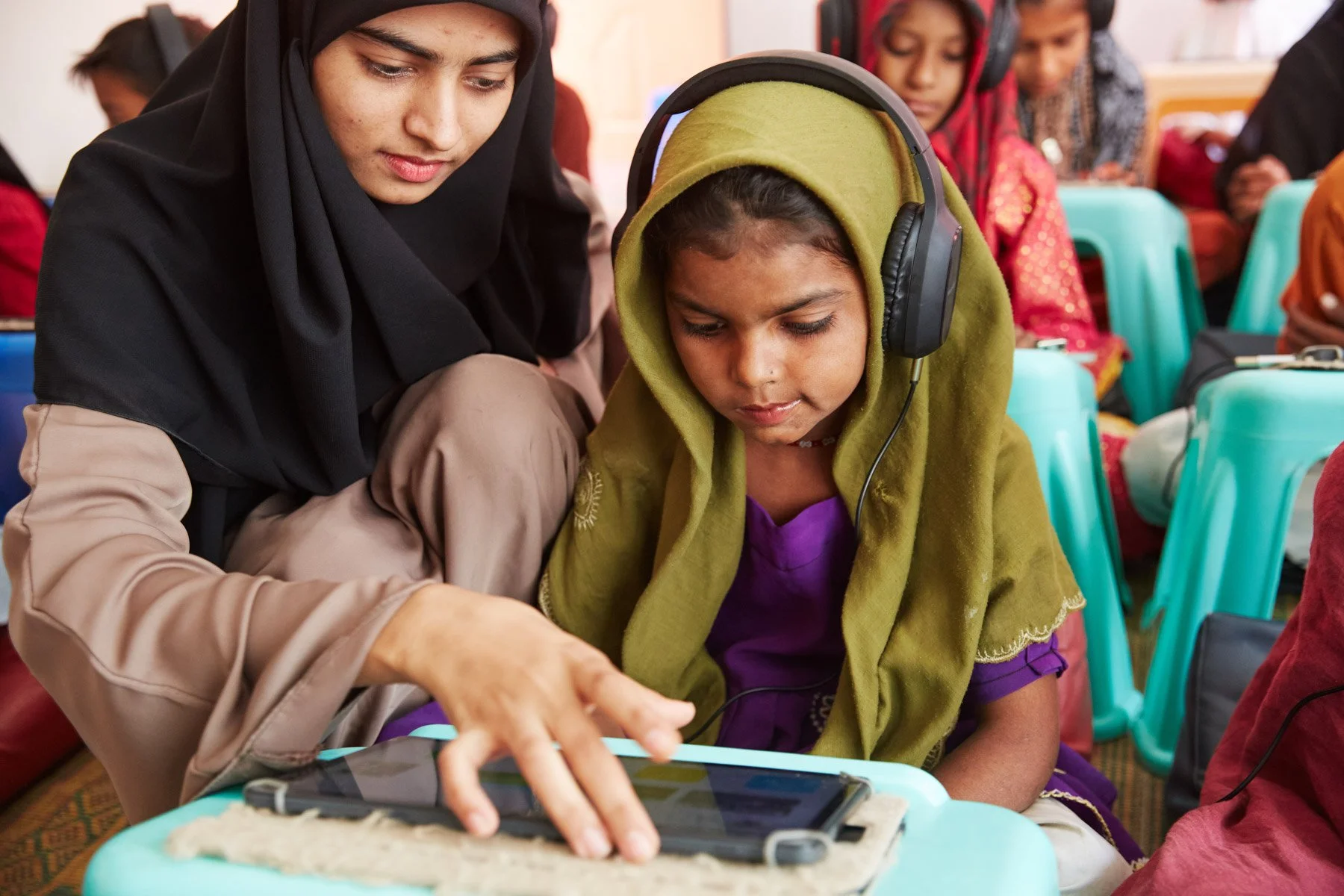 A young girl wearing headphones and a purple dress interacts with a tablet device, guided by an adult woman in a black hijab, in a classroom setting with other children. For Teach the World Foundation, Razzaqabad, Karachi, Pakistan.