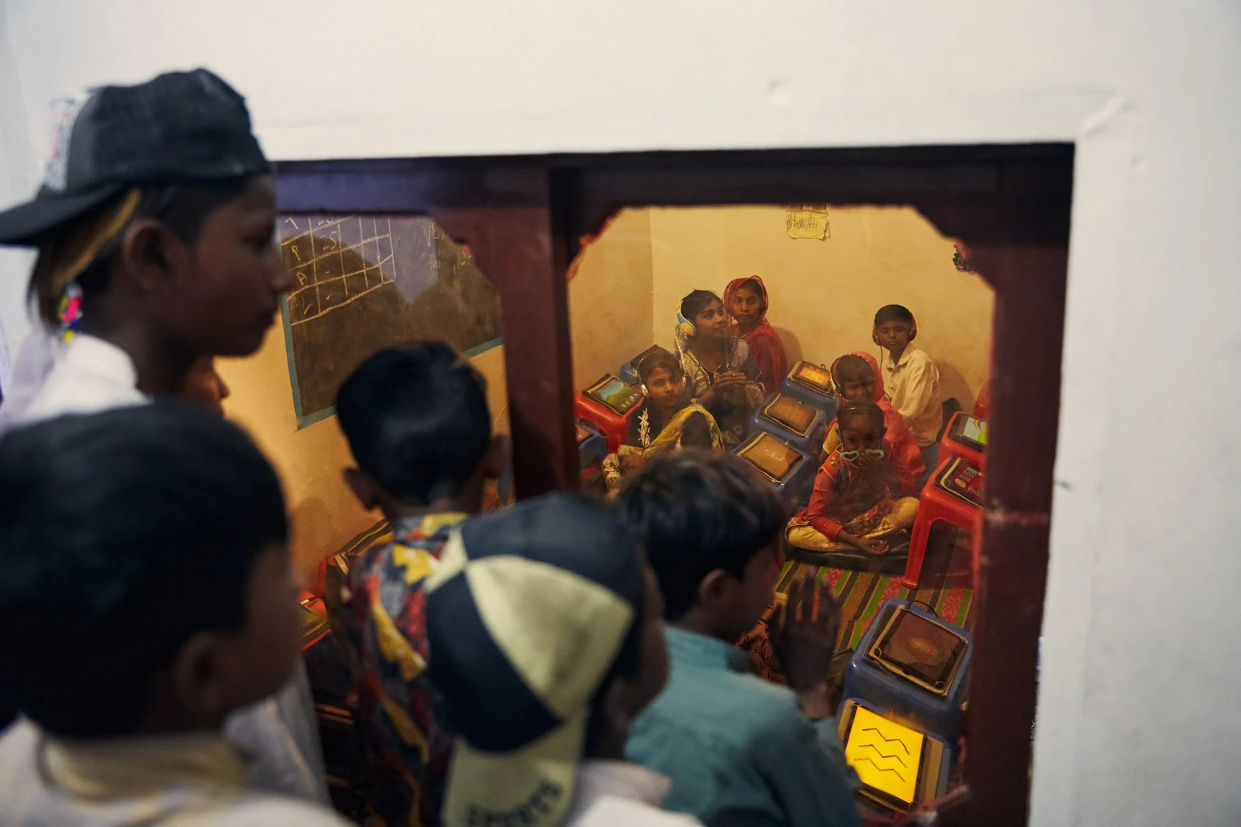Children gathered in a classroom, viewed through a small window. Some children are looking outside, while others are seated at individual tablets. For Teach the World Foundation, Razzaqabad, Karachi, Pakistan.