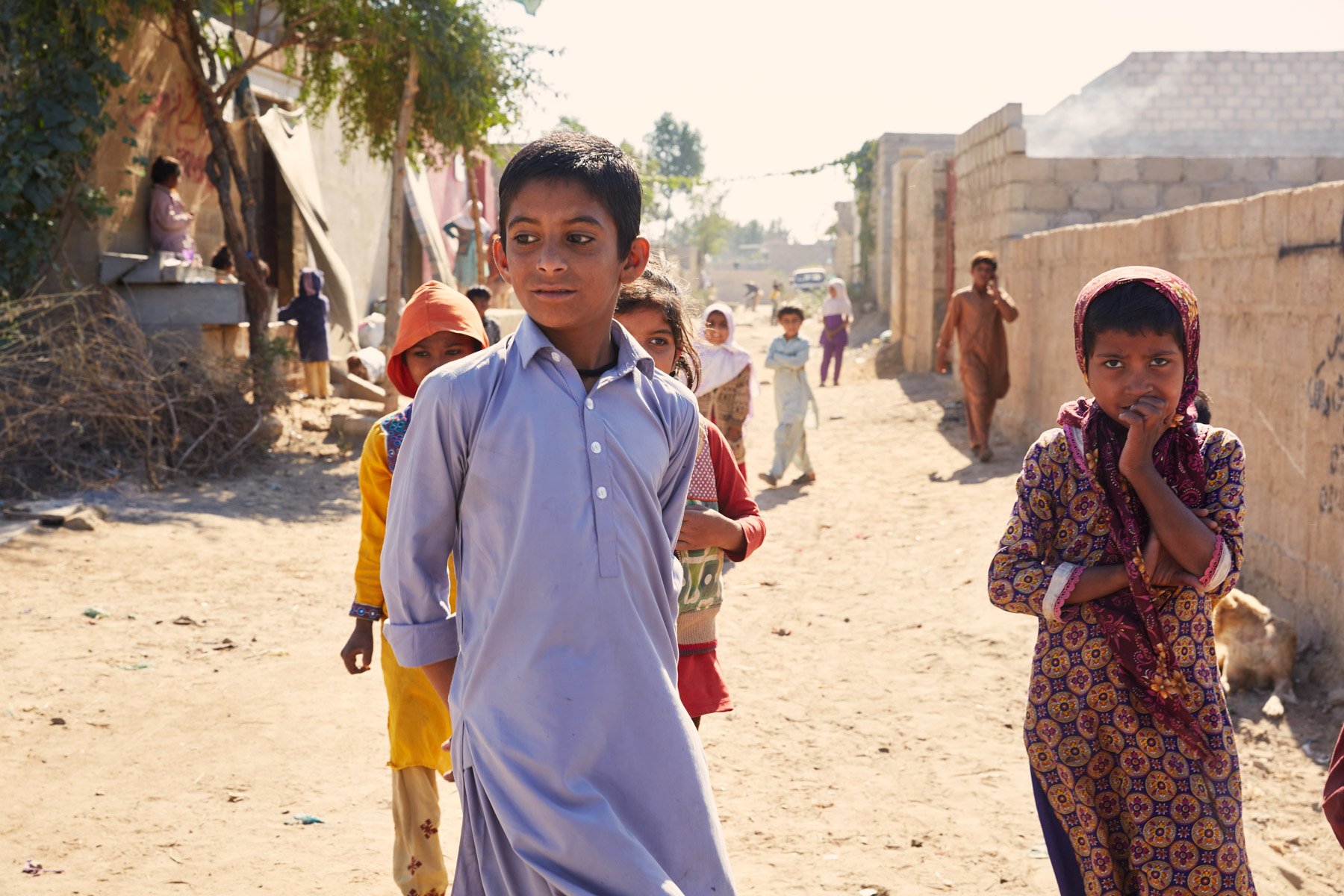 Children walking on a dusty street in Razzaqabad, Karachi, Pakistan. Some children are looking at the camera, while others are engaged in their own activities. The background shows unfinished buildings and a few adults. For Teach the World Foundation