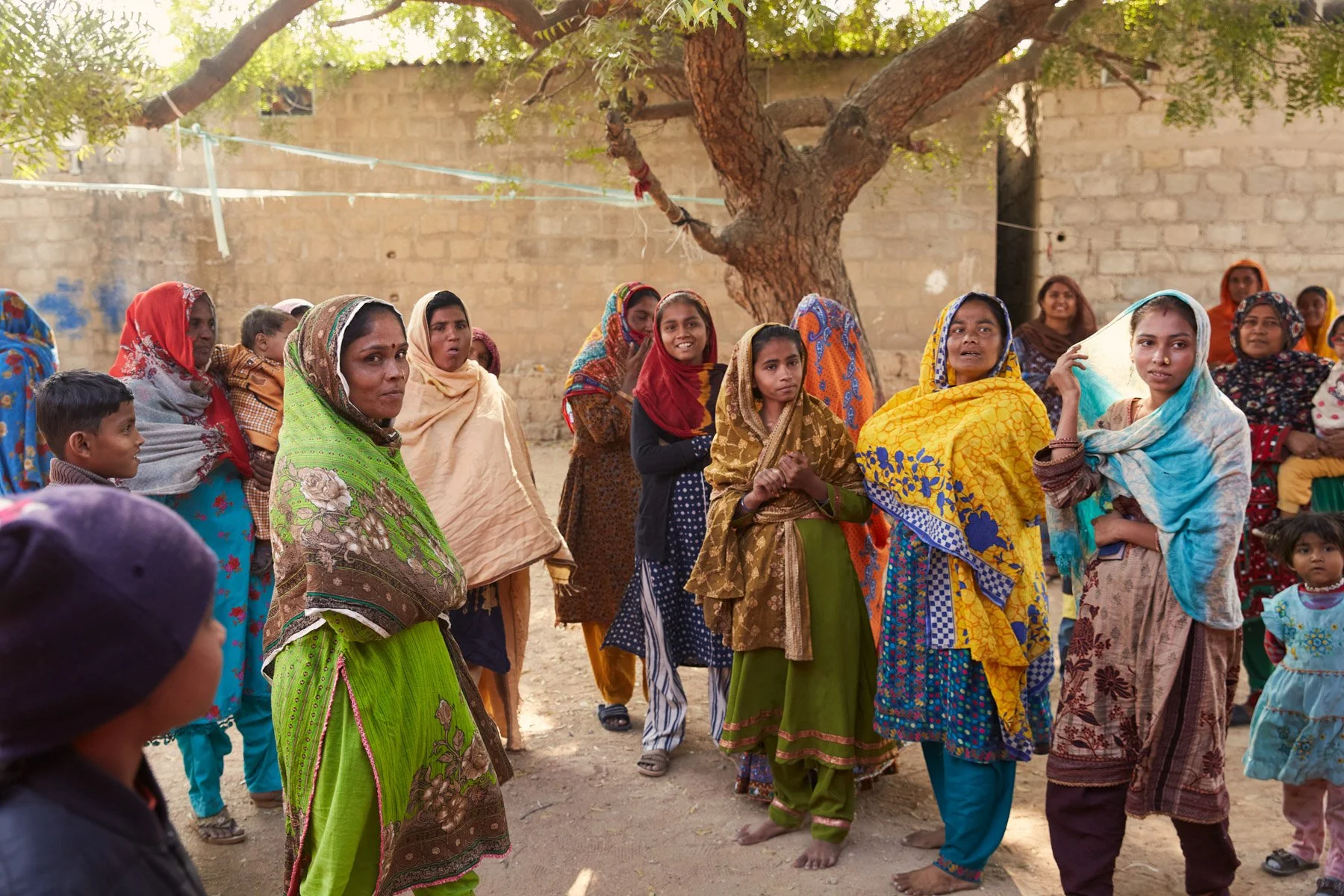 A group of women and children gathered outdoors under a large tree, some wearing colorful traditional clothing and headscarves, with a brick wall in the background. For Teach the World Foundation, Razzaqabad, Karachi, Pakistan.
