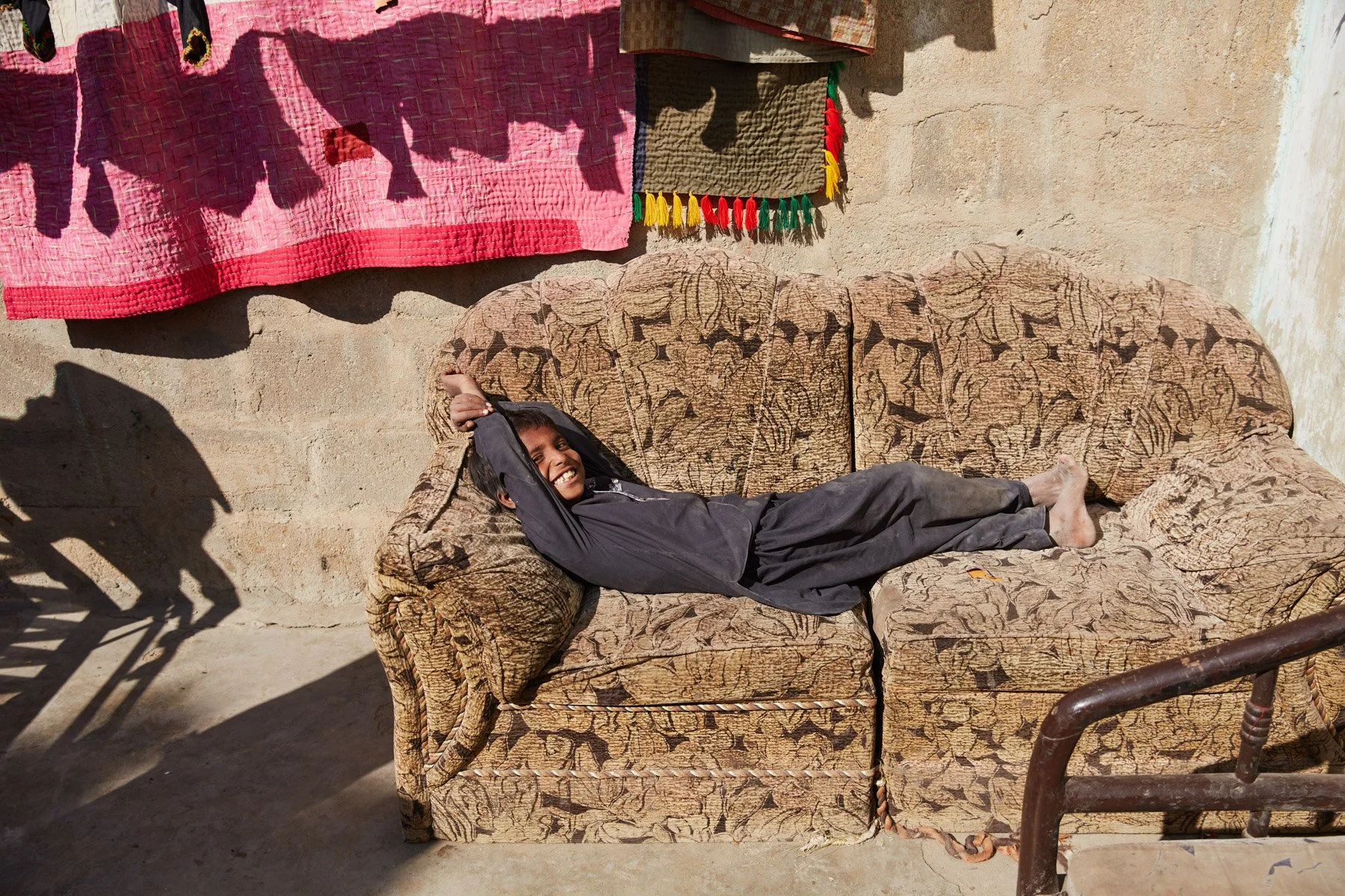 A smiling young boy lying comfortably on a worn, patterned sofa outside against a beige wall with colorful cloth hanging above. Shadows cast by nearby objects are visible on the ground. For Teach the World Foundation, Razzaqabad, Karachi, Pakistan.