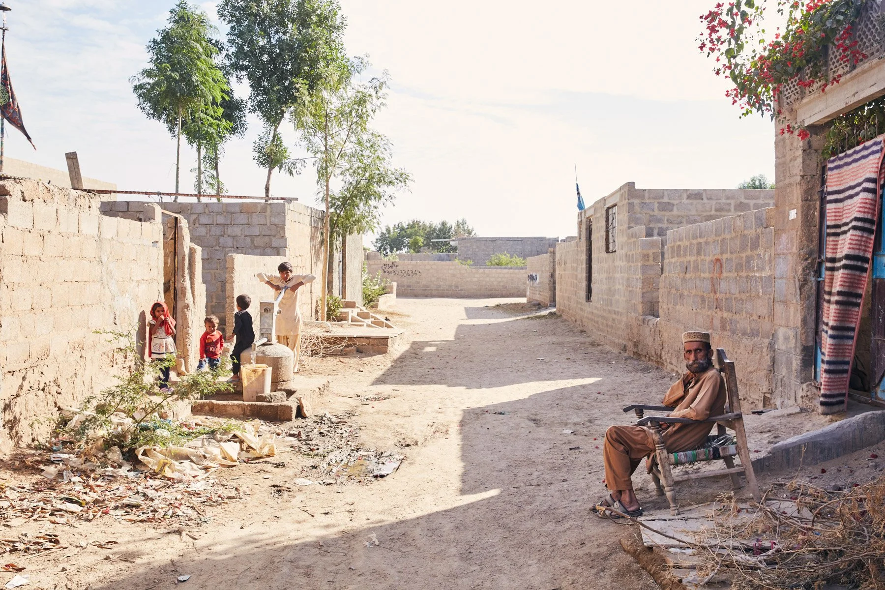 Man sitting on a wooden chair with a book, wearing a traditional cap, on a dirt street with stone houses and children playing nearby. For Teach the World Foundation, Razzaqabad, Karachi, Pakistan.