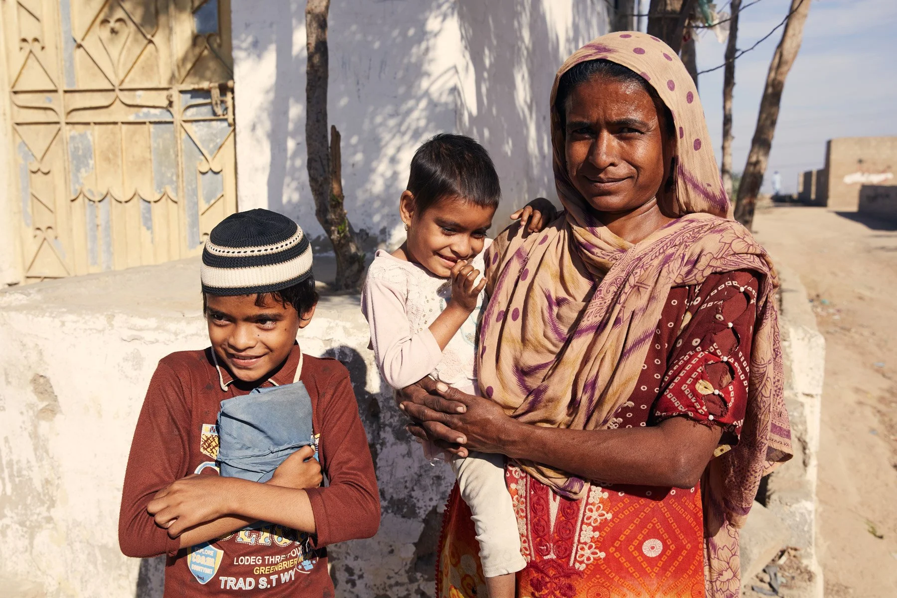 A woman holding a young girl with a smile, standing next to a boy with a tan complexion and a cap, in front of a white wall and a tree, in a rural setting. For Teach the World Foundation, Razzaqabad, Karachi, Pakistan.