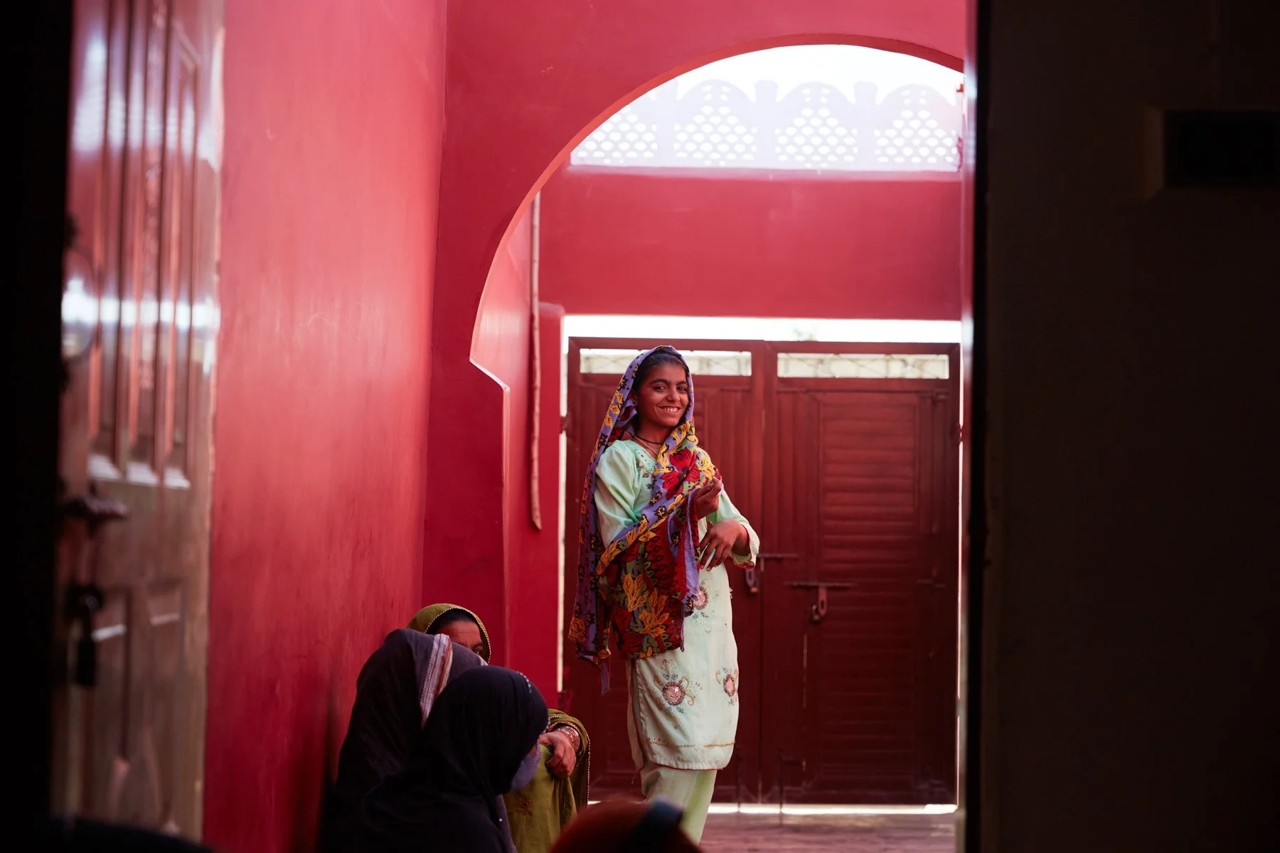 A young woman in traditional attire smiling and standing in a doorway with a red wall and gate behind her. For Teach the World Foundation, Razzaqabad, Karachi, Pakistan.