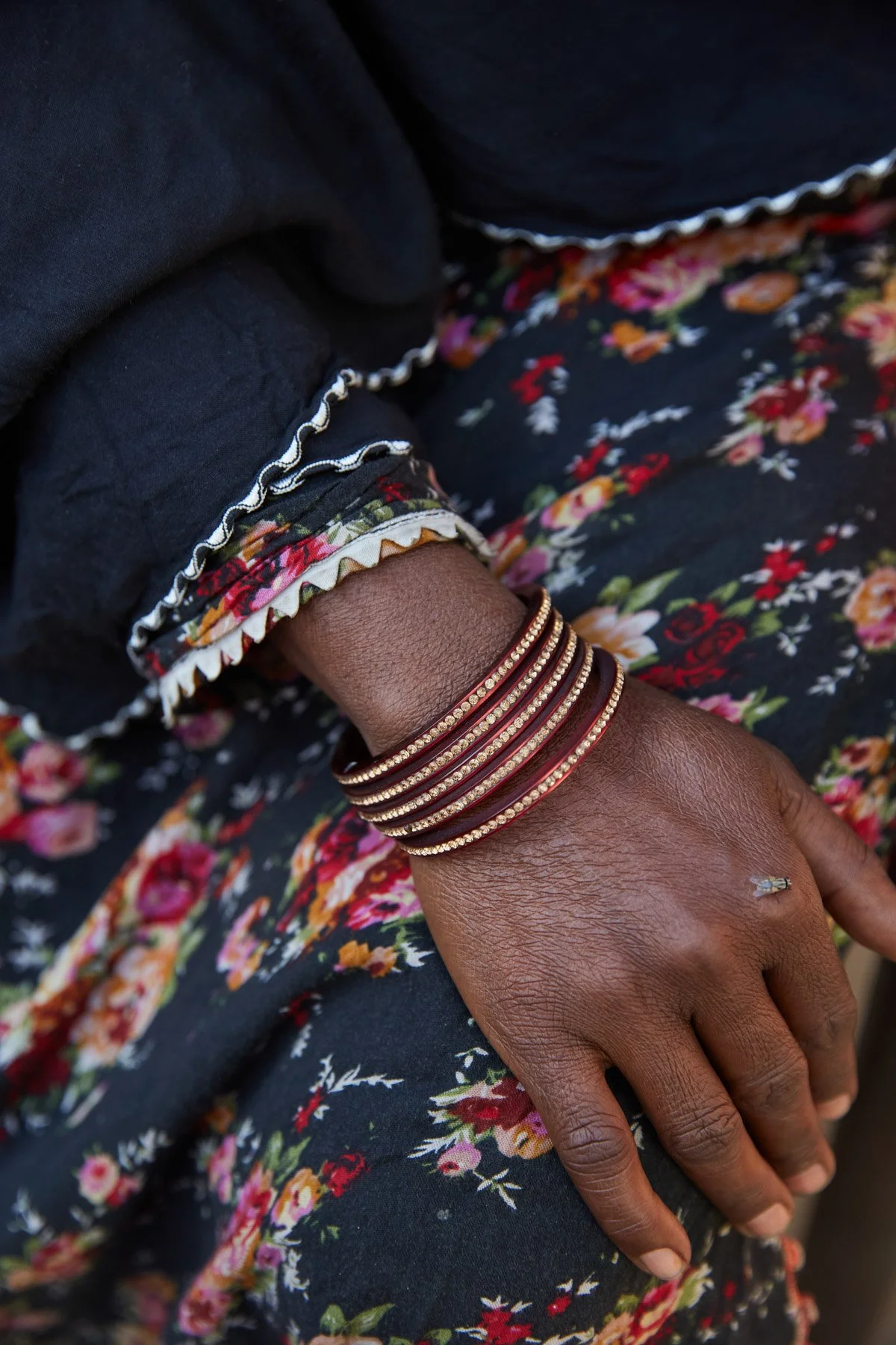A woman's hand resting on her lap, adorned with multiple gold bangles, wearing a floral dress and a black top with embroidered trim. For Teach the World Foundation, Razzaqabad, Karachi, Pakistan.