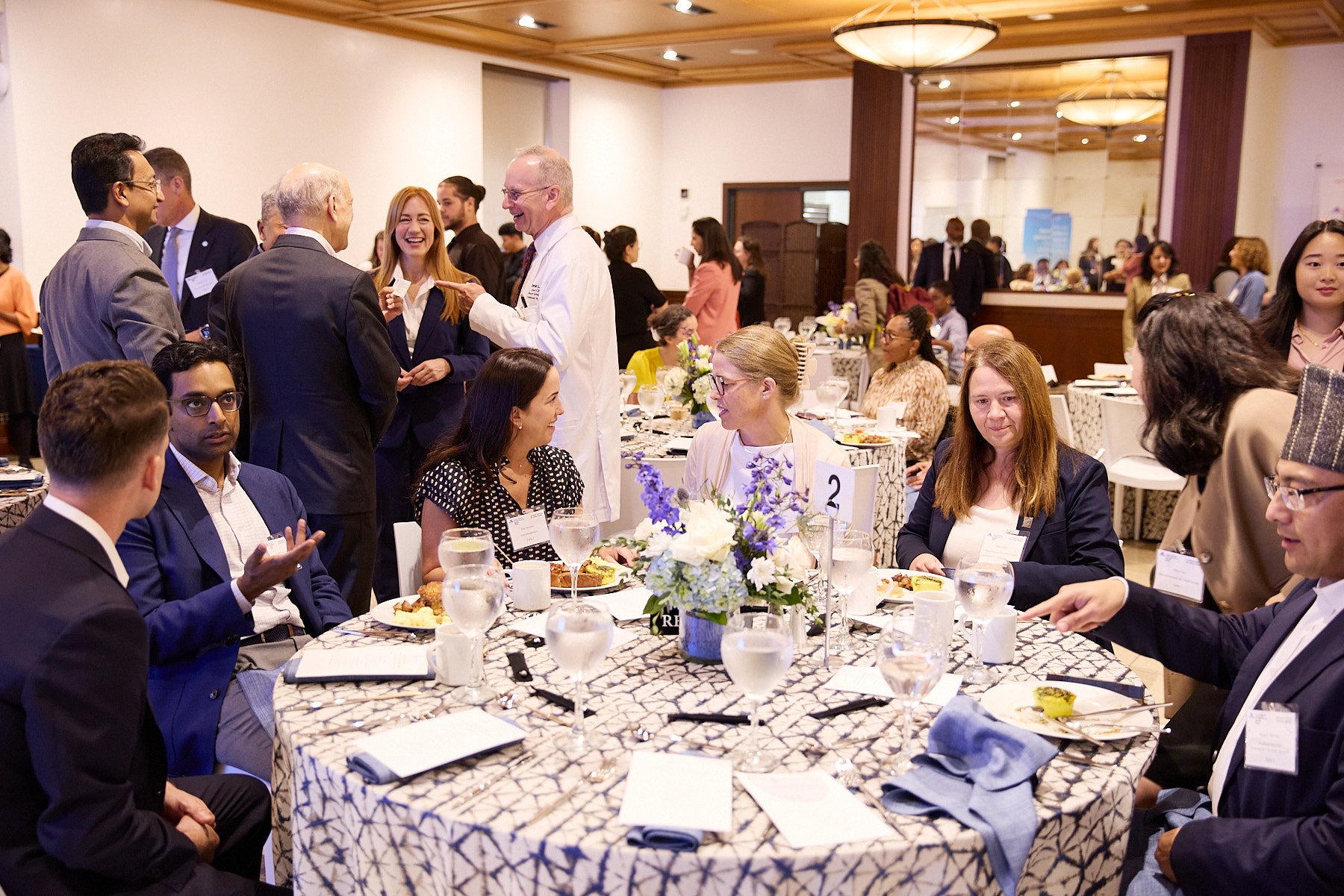 A diverse group of people at a formal event, some seated at a round table with floral centerpiece, others standing and chatting in a banquet hall.