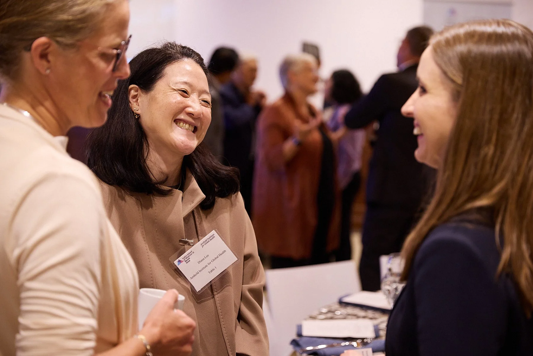 Three women engaging in a lively conversation, smiling and laughing at a professional event or conference, with people in the background.