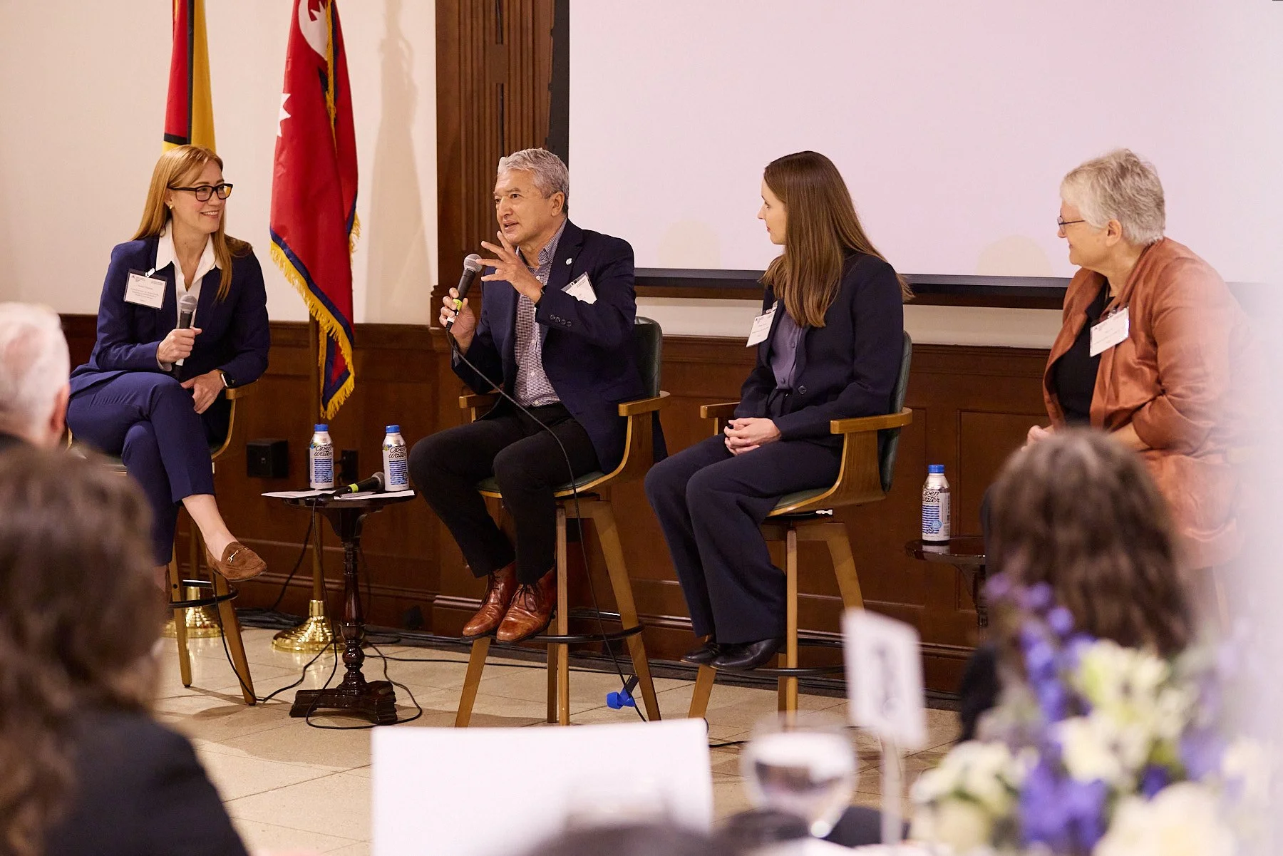 Four women and one man sitting on stage in a panel discussion. The man is speaking into a microphone. There are flags in the background and bottles of water on small tables.