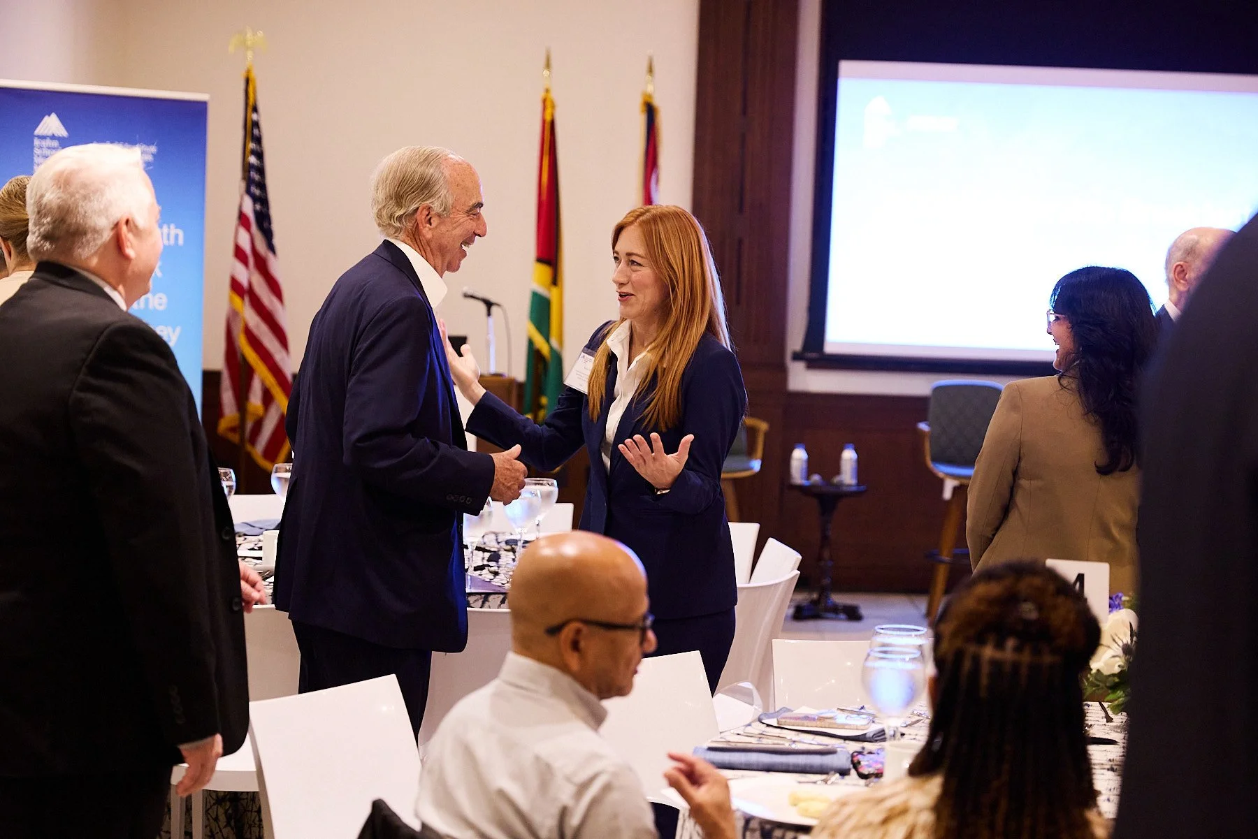 At a professional event, a woman with red hair and a man with gray hair shake hands and talk, with other attendees in the background.
