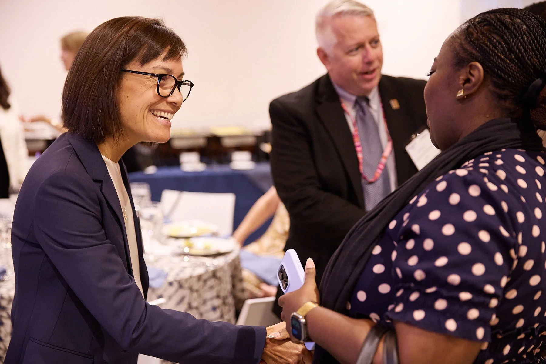 Three people engaged in conversation at a formal event. The woman on the left is smiling and shaking hands with the woman on the right, who is holding a phone. A man in a suit is talking in the background.