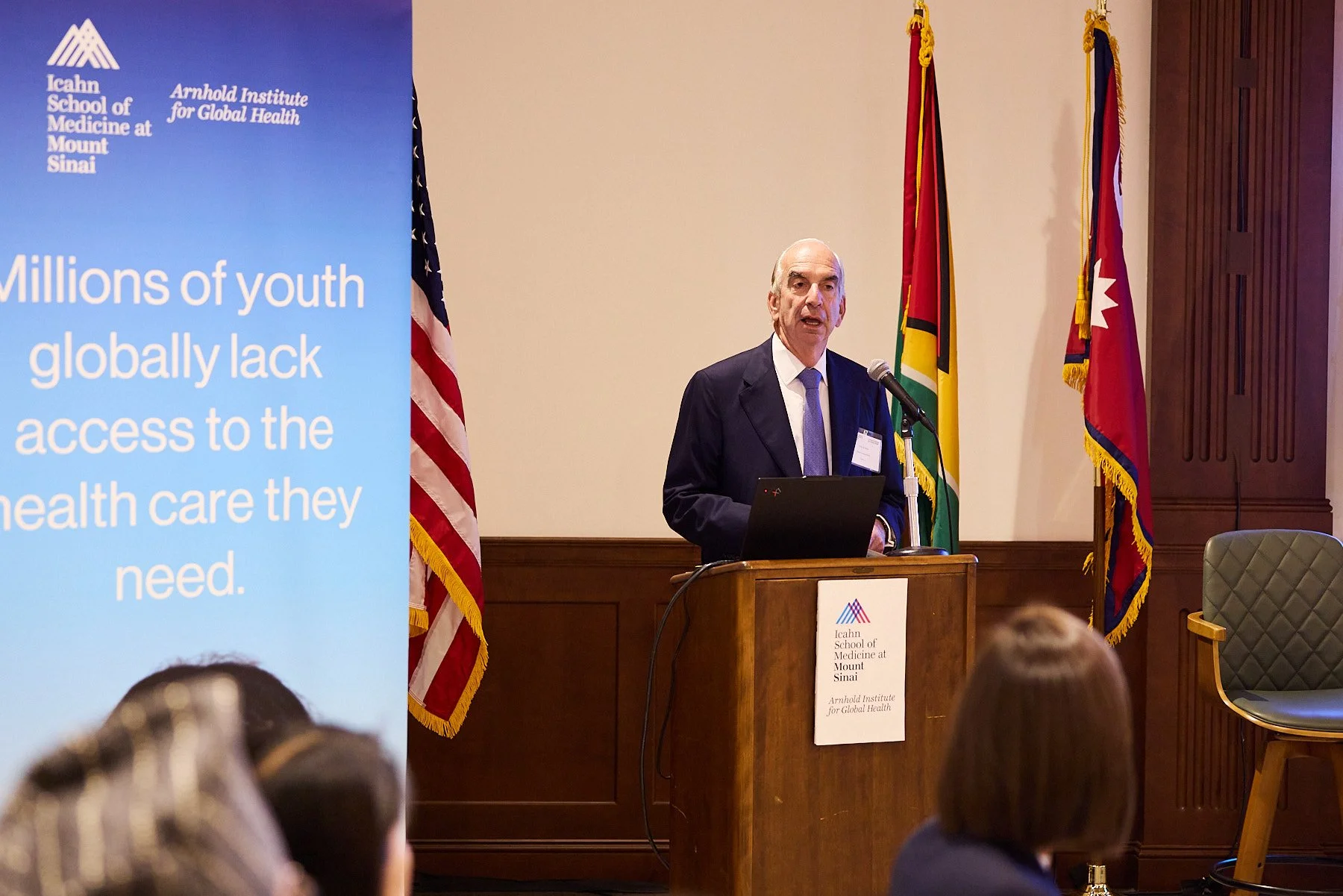 A man in a suit and tie is giving a speech at a podium with a laptop, behind him are several flags including the American flag and others, in a conference room with a large blue presentation screen on the left side displaying text about youth and hea
