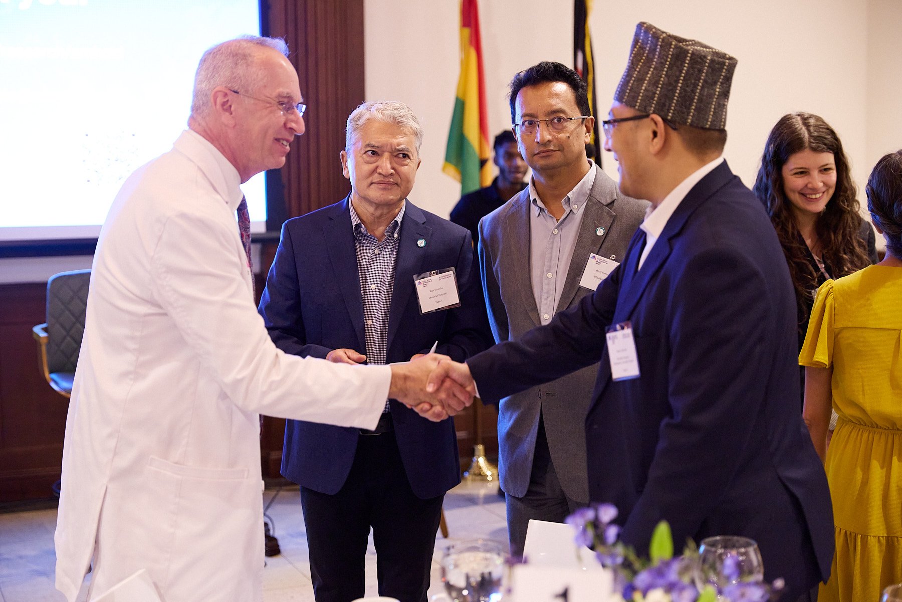 A group of people in formal attire shaking hands at a conference or event, with flags in the background.