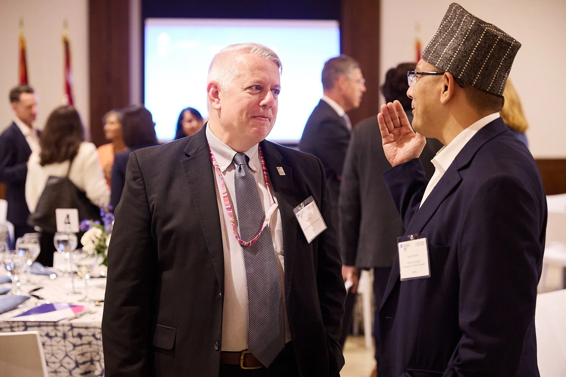 Two men in suits and glasses talking at a professional event, with a background of other attendees, conference room decor, and a large screen.