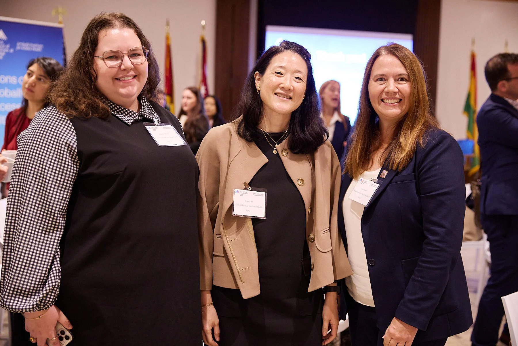 Three women standing together at a conference or event, smiling for the camera. They are wearing business casual attire with name tags, and there are other people and flags in the background.