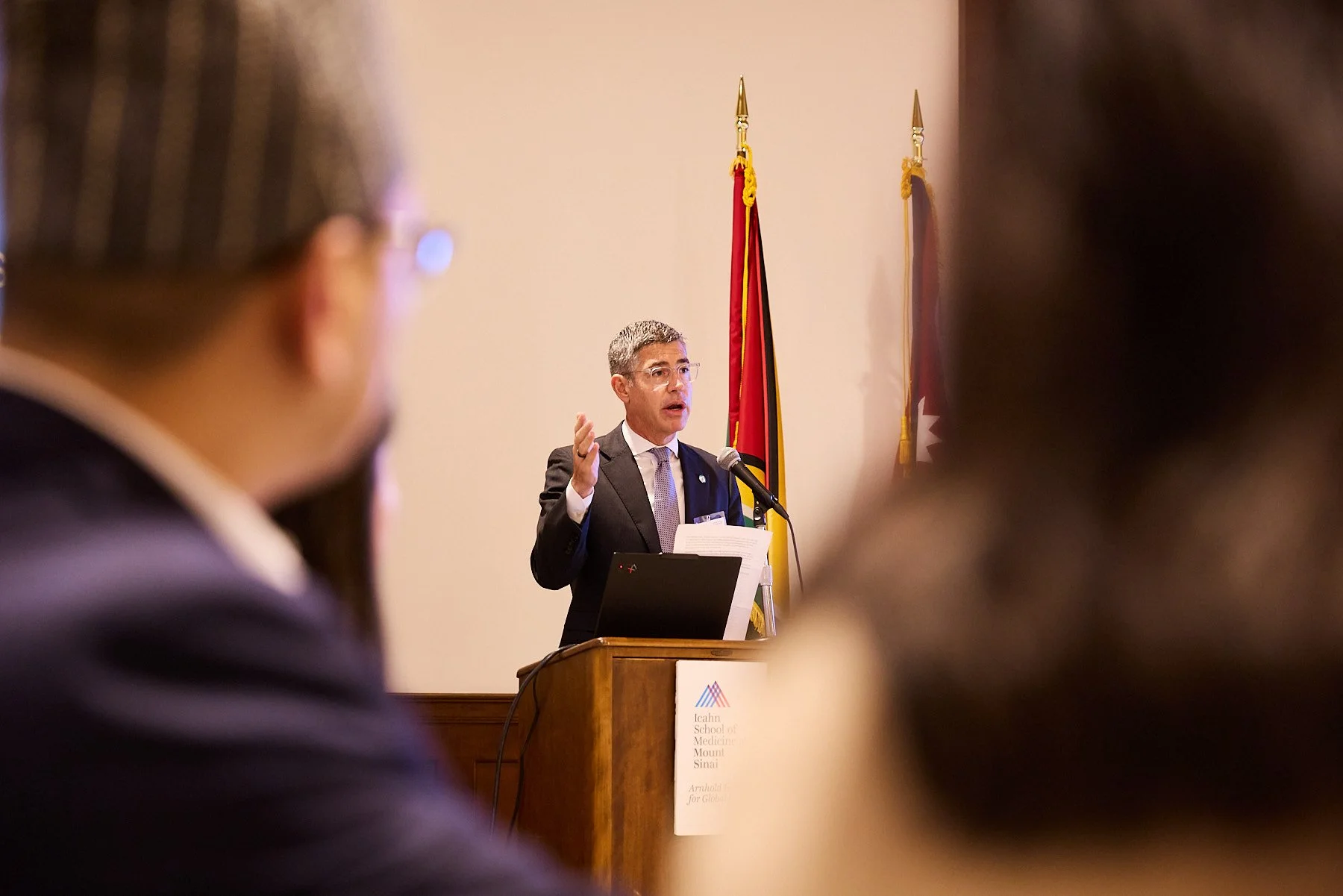 A man in a suit giving a speech at a podium with two flags behind him, observed by an audience.