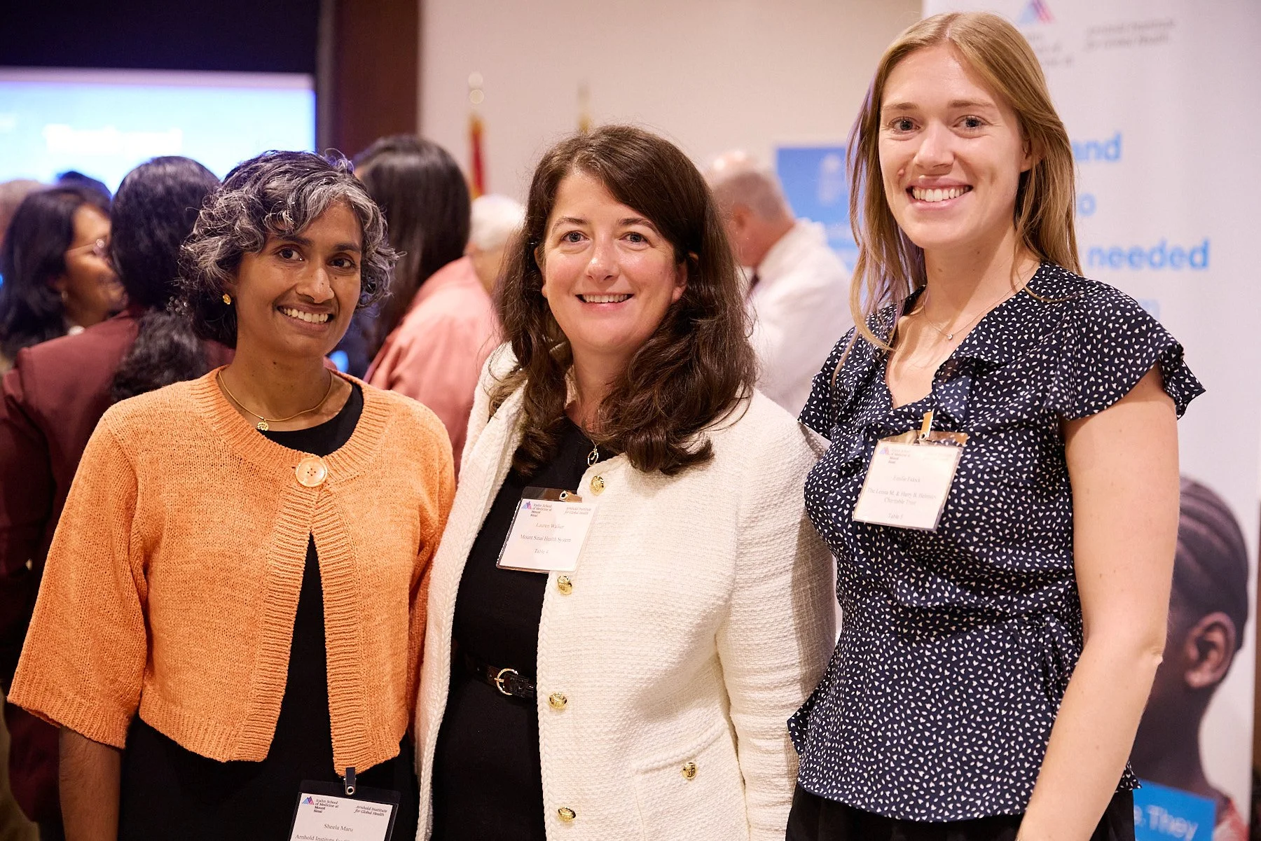 Three women at a conference or event, standing together and smiling at the camera, with name tags and a blurred background of other attendees.