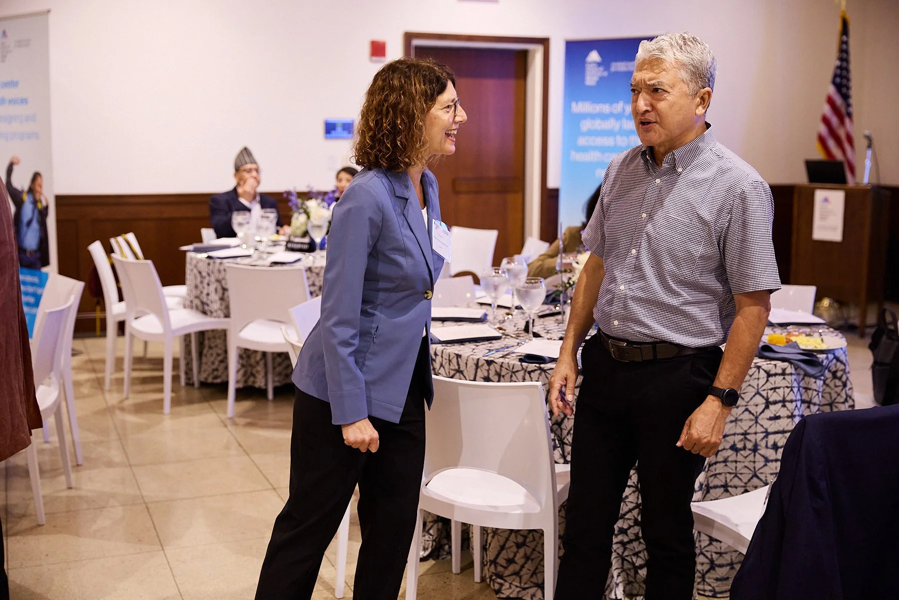 Two people, a woman with curly hair in a blue blazer and a man with gray hair in a checkered shirt, are conversing in a conference room set for a formal event.