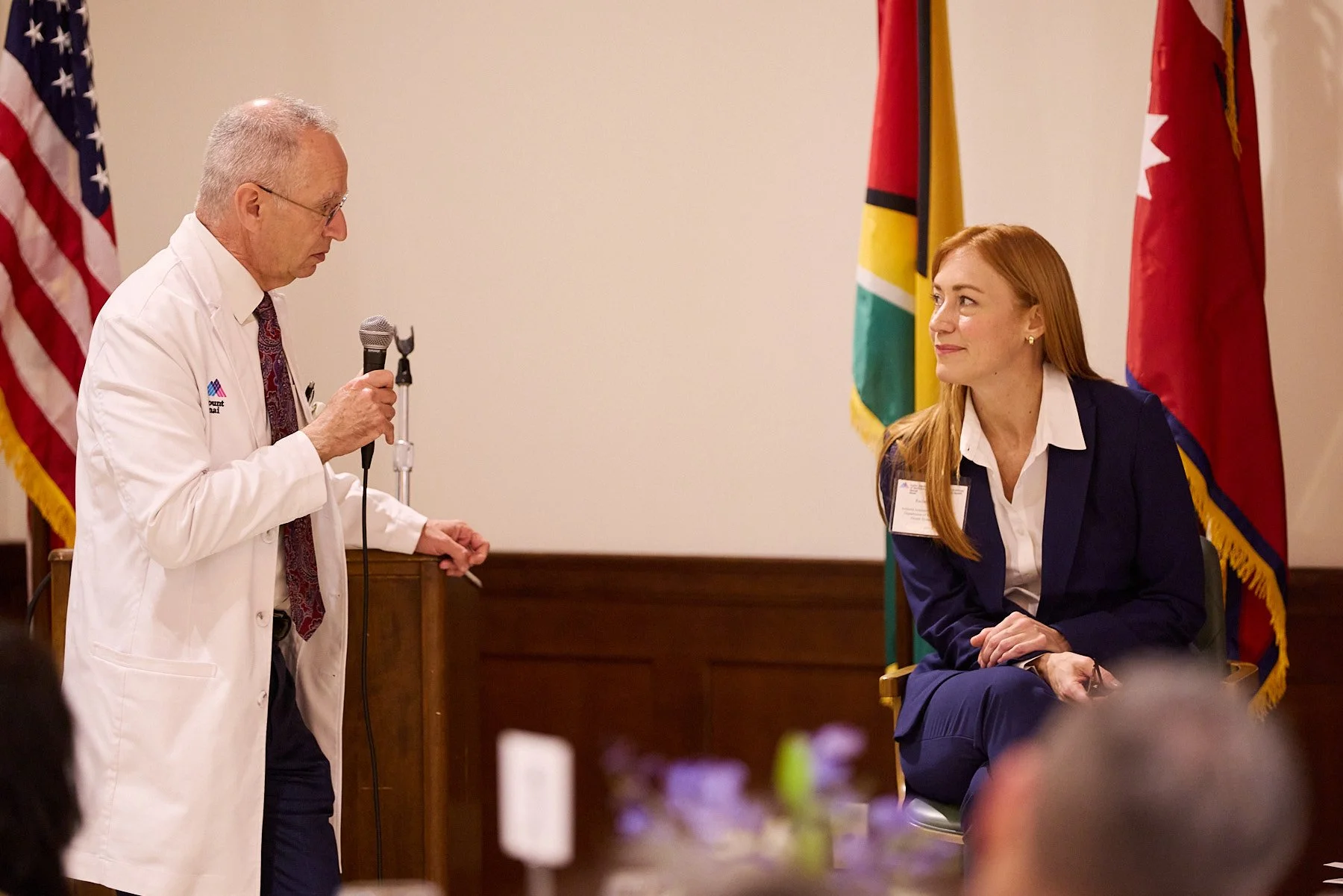 A man in a white coat and glasses holding a microphone is speaking to a woman seated to his right, who is looking at him with a smile. The woman has long red hair and is wearing a navy blazer and white shirt. They are in a room decorated with America