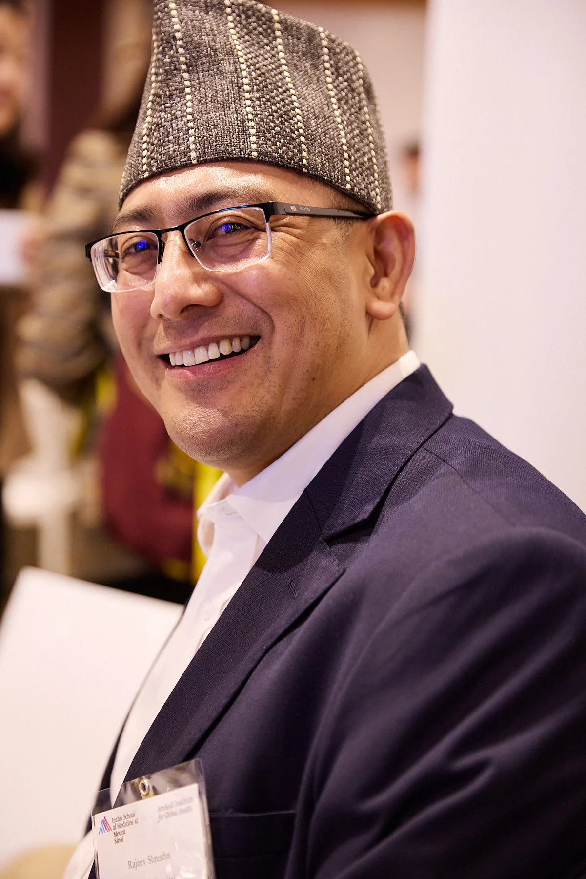 A man wearing glasses, a patterned traditional hat, a white shirt, and a dark suit blazer, smiling at a professional or formal event.