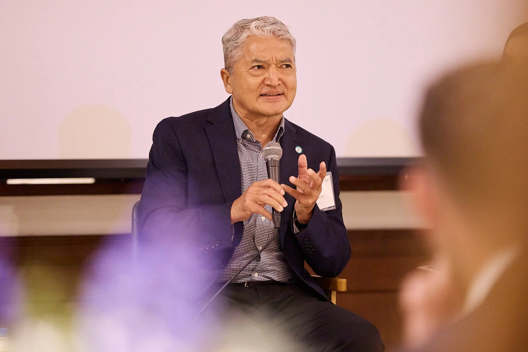 An older man with gray hair, wearing a dark blazer and a checkered shirt, is sitting and speaking into a handheld microphone during a presentation or panel discussion.