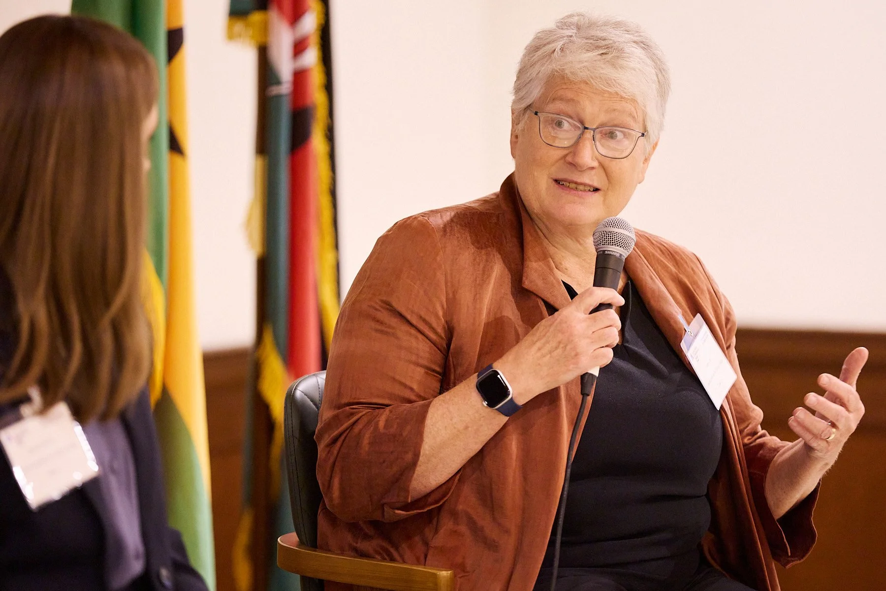 An older woman with short gray hair and glasses, wearing a black top and a brown jacket, is speaking into a microphone during a panel discussion. She is gesturing with her left hand, and a smartwatch is visible on her left wrist. Behind her, there ar