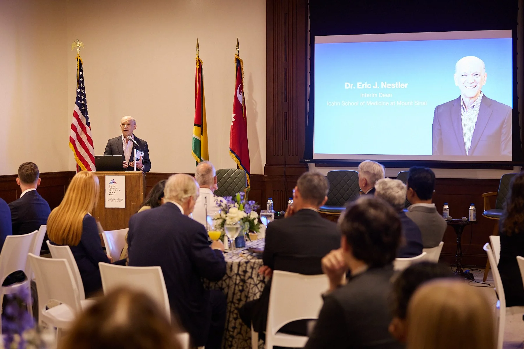 A man giving a presentation at a conference, standing at a podium with a laptop, with the American, Kenyan, and another national flag behind him. A large screen displays a slide with the name Dr. Eric J. Nestler and his title. Attendees are seated at