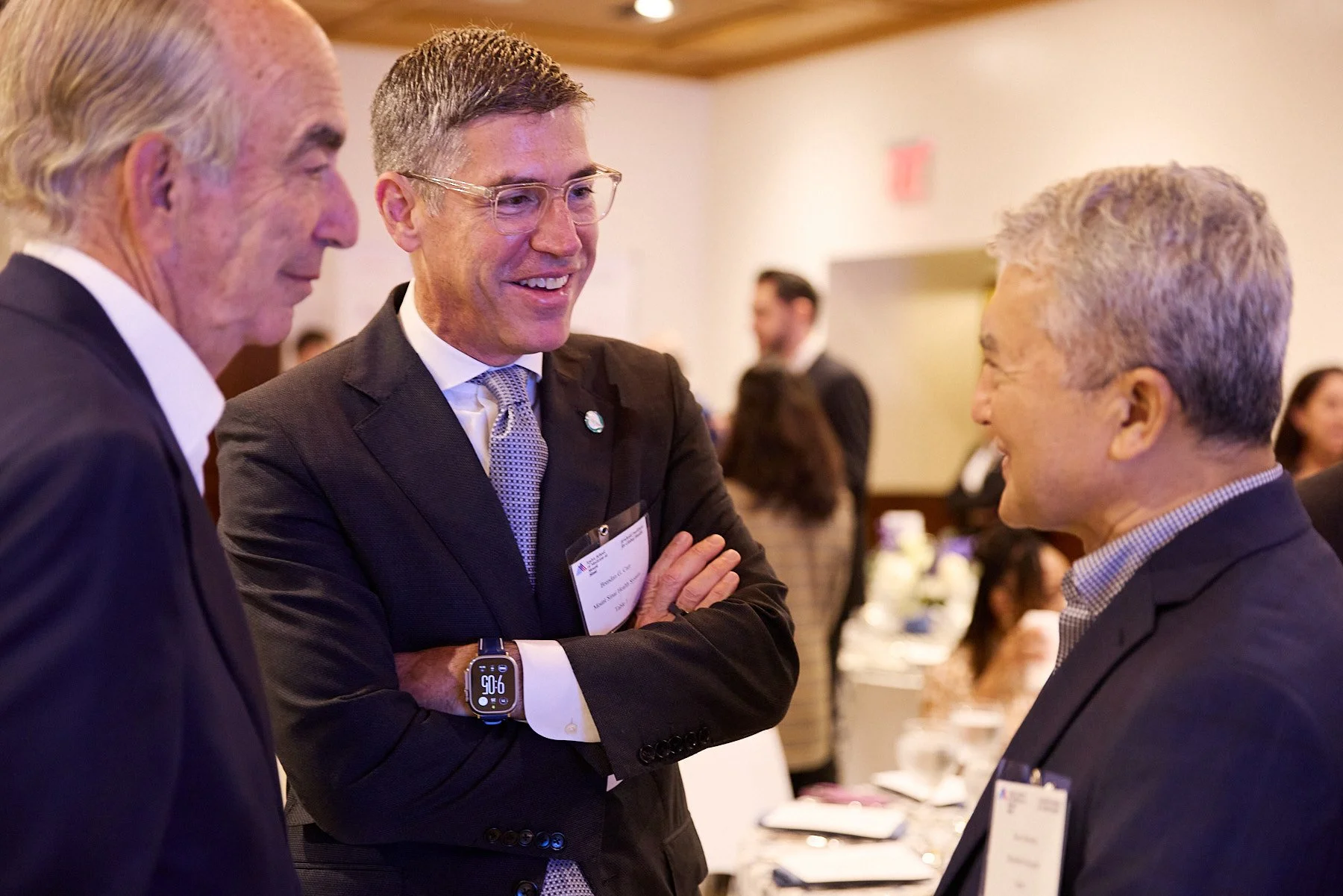Three men engaged in conversation at a professional event. They are wearing suits and conference name tags. The background shows other attendees and a formal dining setup.
