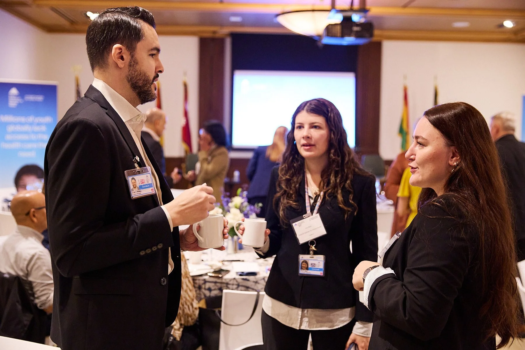 Three business professionals with name badges and coffee cups, engaged in conversation at a conference or networking event in a formal setting.