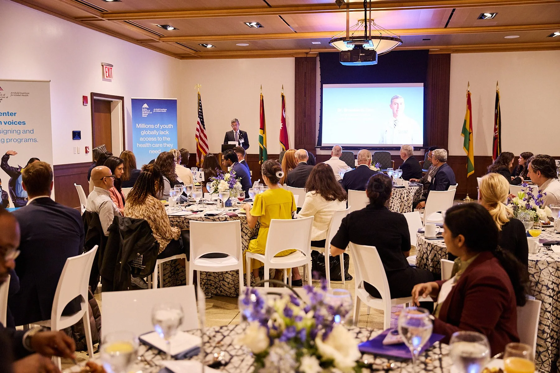 Conference with a speaker on stage and attendees seated at round tables with floral centerpieces, in a room decorated withFlags and a large screen displayed.