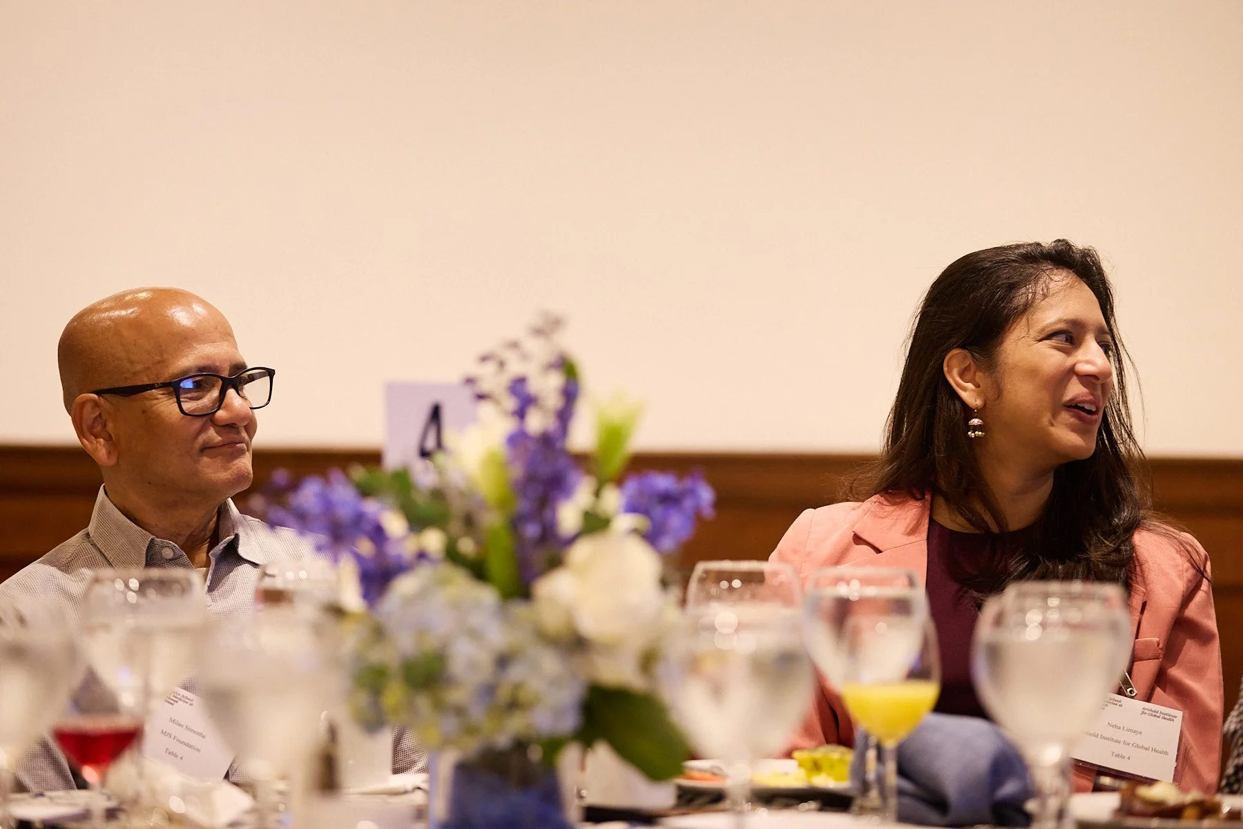 A man with glasses and a woman sitting at a table during a formal event with glasses and a floral centerpiece.