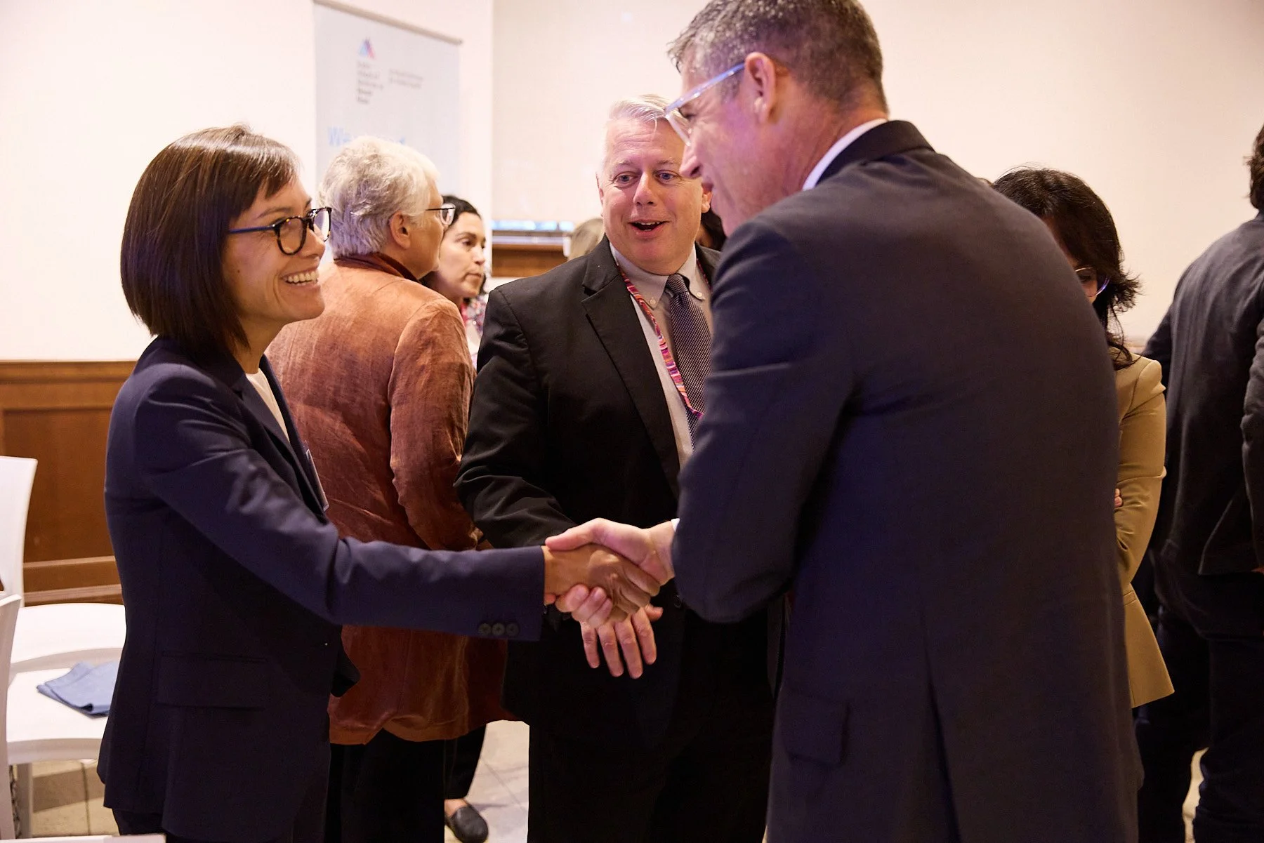 A woman in a navy blazer and glasses shaking hands with a man in glasses and a dark suit in a professional setting, with other people engaging in conversation in the background.