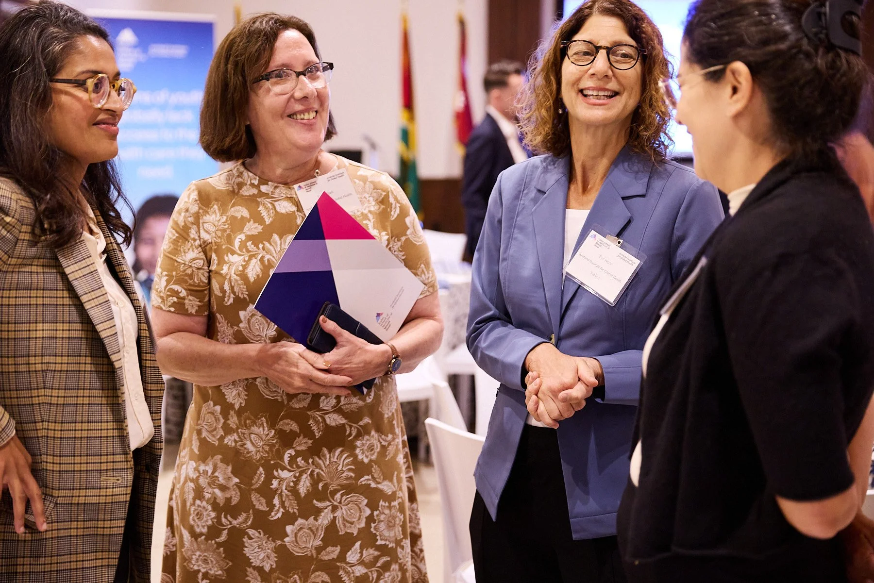 Four women at a professional conference or event, engaged in conversation. One woman is shaking hands with another, and they are all smiling.