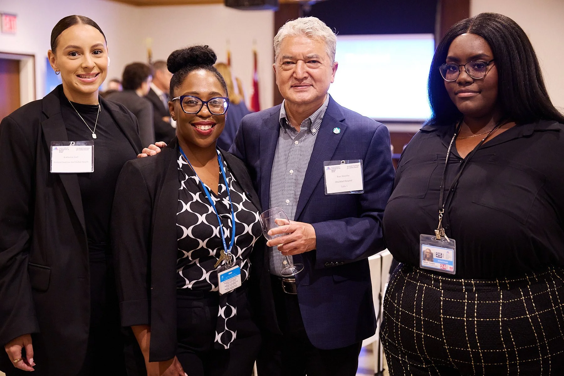 Four people at a professional event posing for a photo, three women and one man, all wearing business attire and name badges.