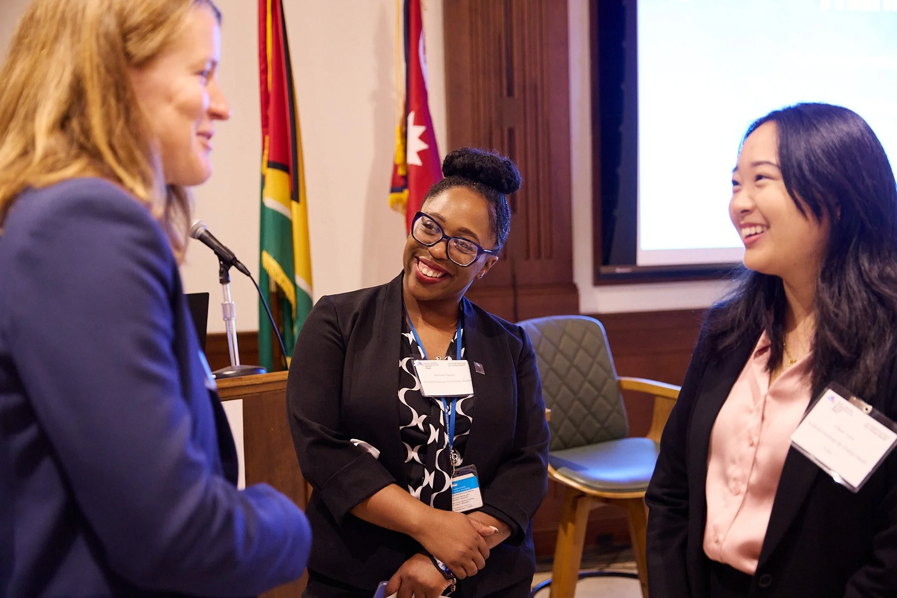 Three women in business attire smiling and talking in a conference room, with flags and a screen in the background.