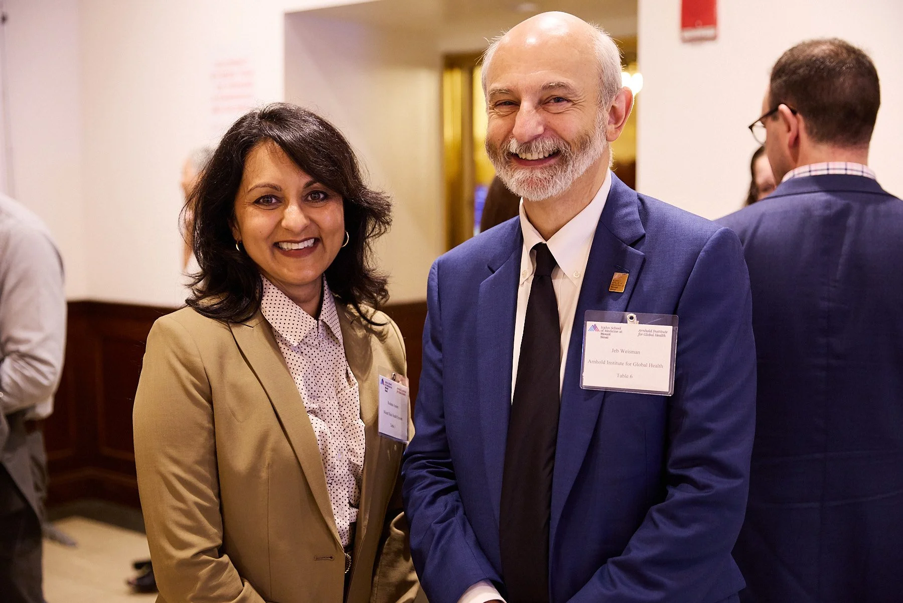 Two professionals, a woman and a man, smiling at a formal event. The woman has dark hair and is wearing a tan blazer over a polka dot blouse. The man has a beard and is dressed in a blue suit with a black tie. Both are wearing conference name tags.