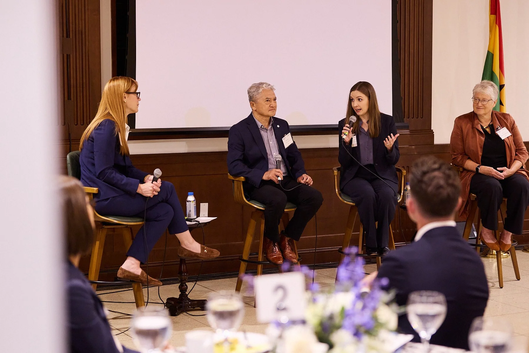 Four women are sitting on chairs on a stage, participating in a panel discussion at an event. One woman is speaking into a microphone, while the others listen. There are tables with water glasses and flowers in the foreground, and a large white scree