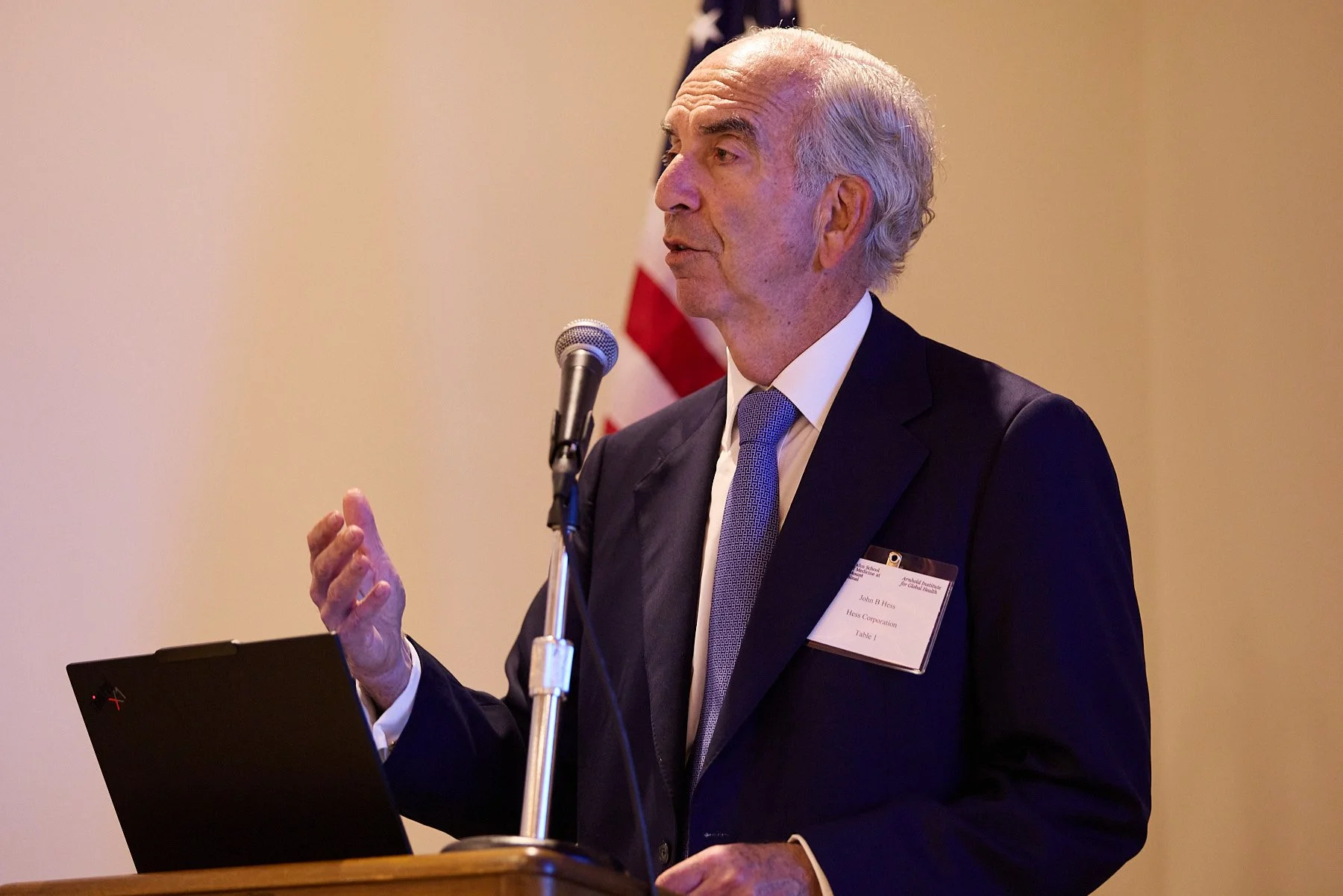 An older man with gray hair in a suit and tie is speaking at a podium with a microphone, gesturing with his hand. There is an American flag in the background.
