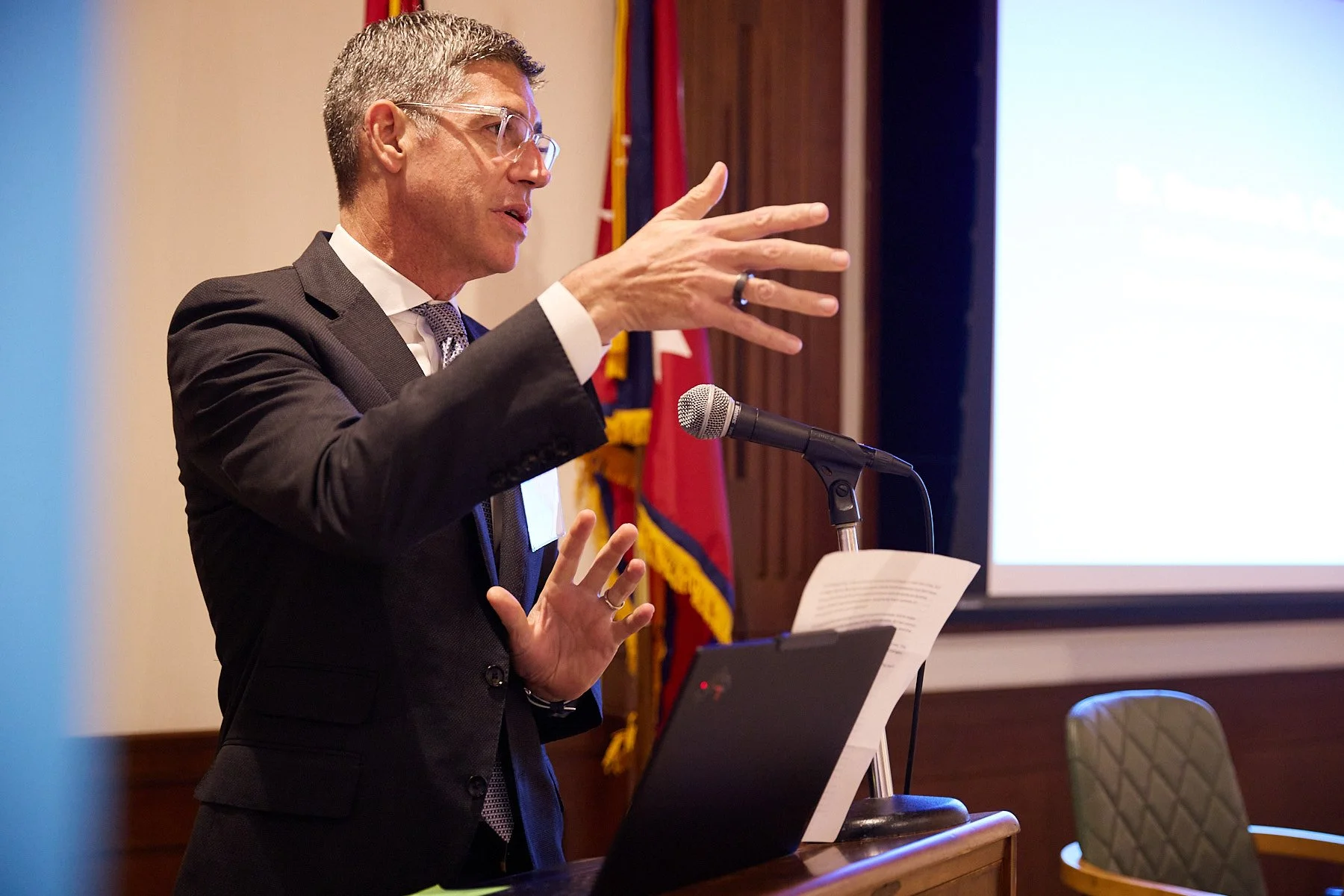 A man in a suit and glasses is speaking at a podium in front of a microphone, gesturing with one hand, with a laptop and papers on the podium, and a presentation screen in the background.