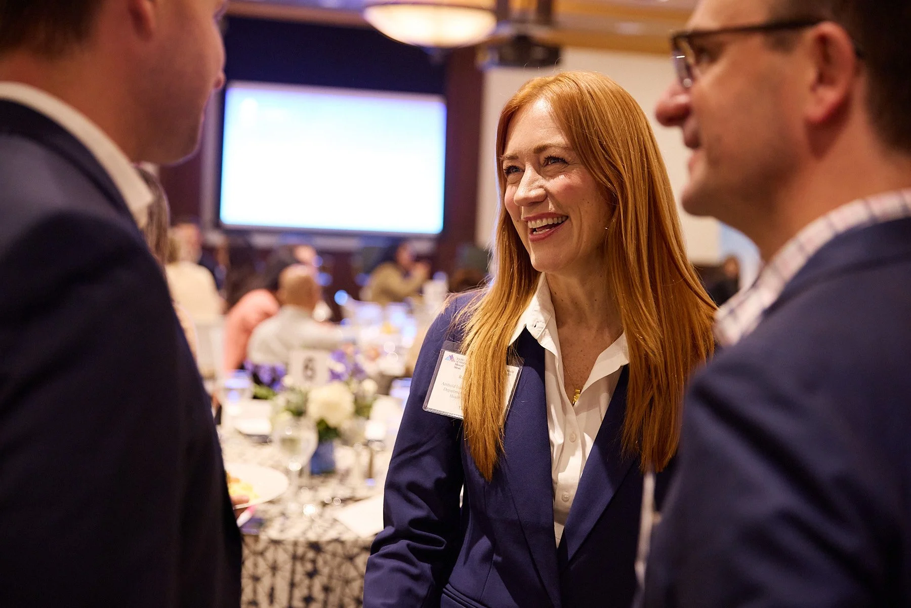 A woman with long red hair, wearing a dark blue blazer and white shirt, is smiling while talking to two men at a formal event or conference.
