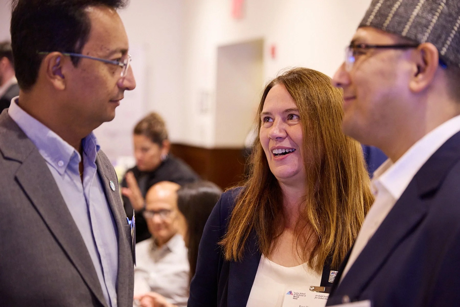 Three people engaged in conversation at a professional event or conference, with other attendees visible in the background.
