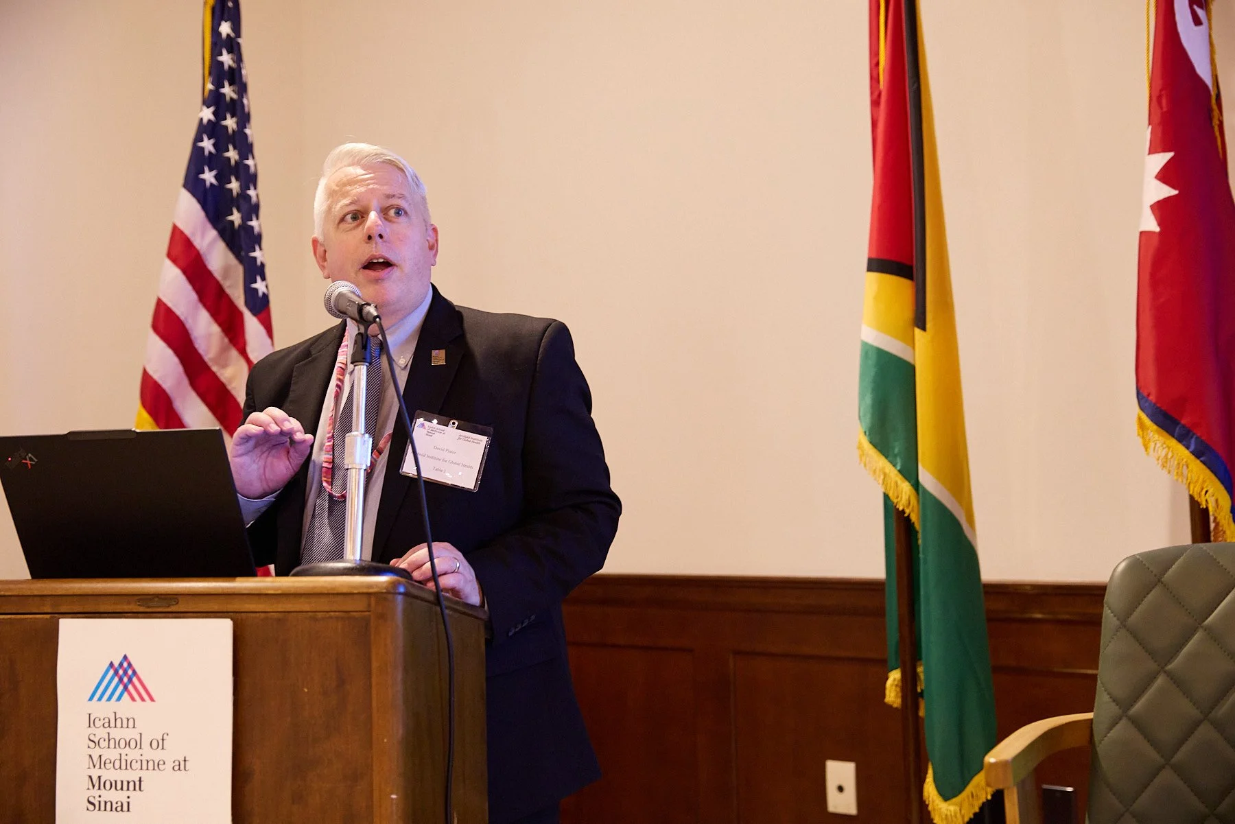 A man in a black suit speaking at a podium during a conference or presentation with American and other flags behind him. The podium has a sign that reads 'Icahn School of Medicine at Mount Sinai'.