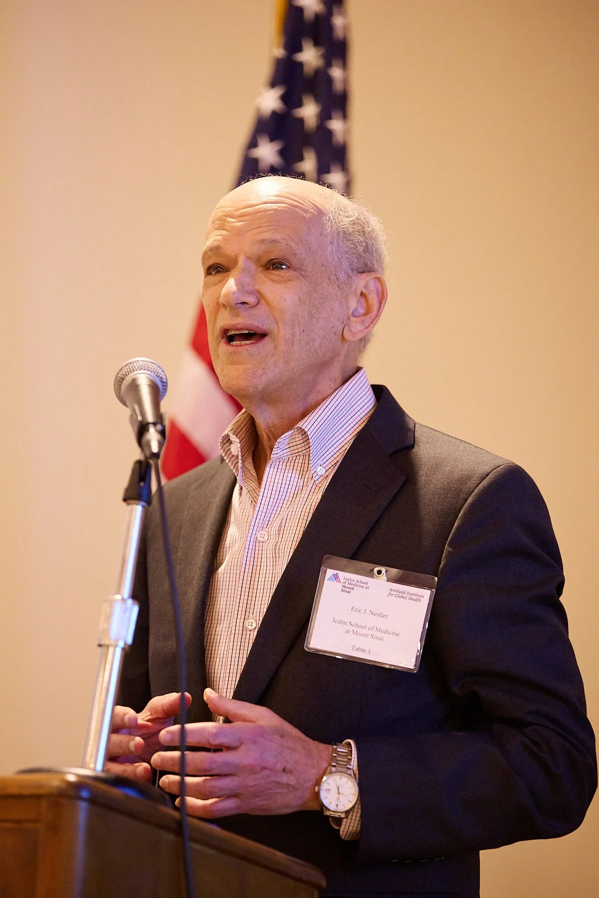 An older man speaking at a podium during a formal event, wearing a dark suit, a light checked shirt, a wristwatch, and a name badge. An American flag is visible in the background.