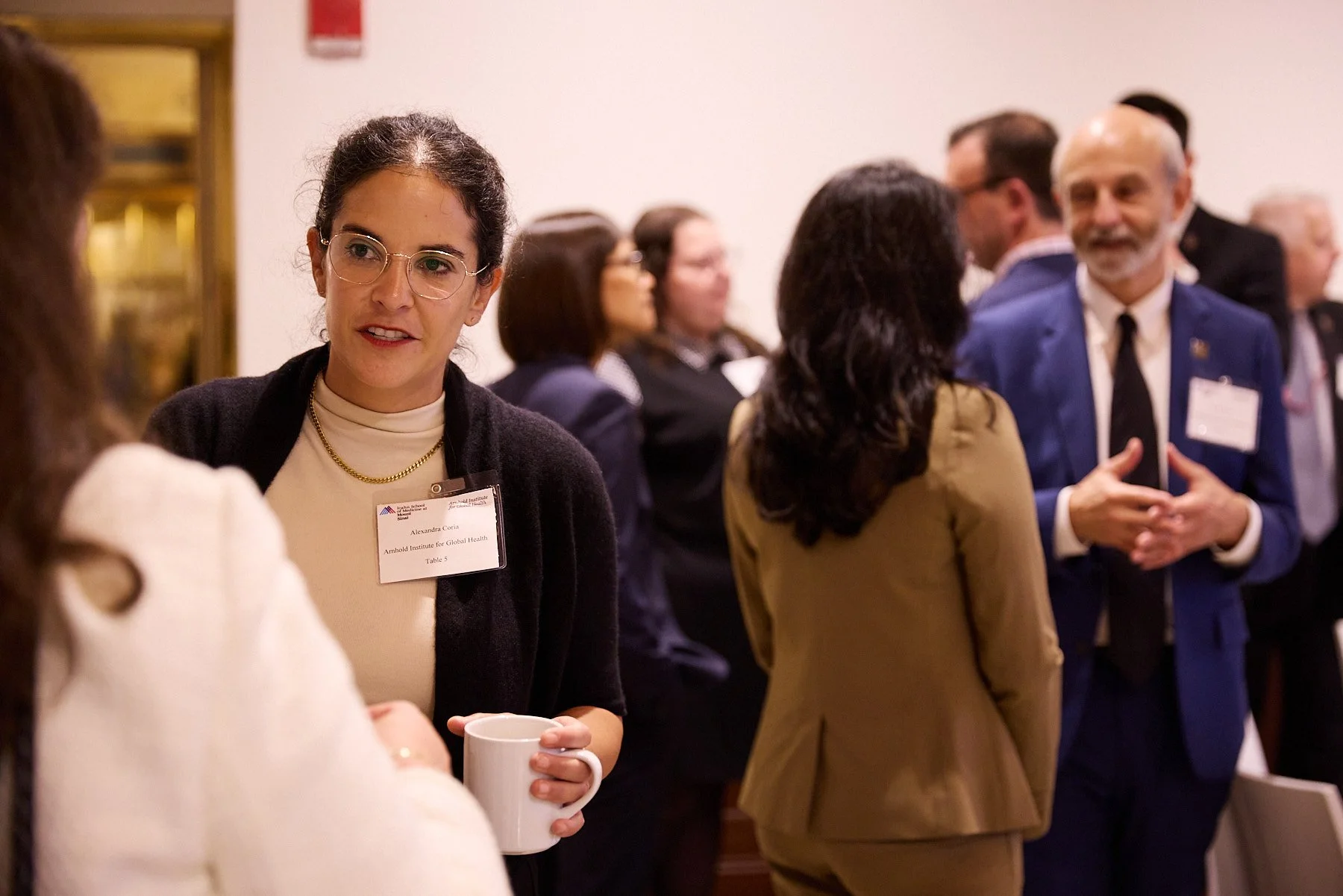 Conference or networking event with people engaging in conversations, including a woman with glasses holding a mug and wearing a name tag.