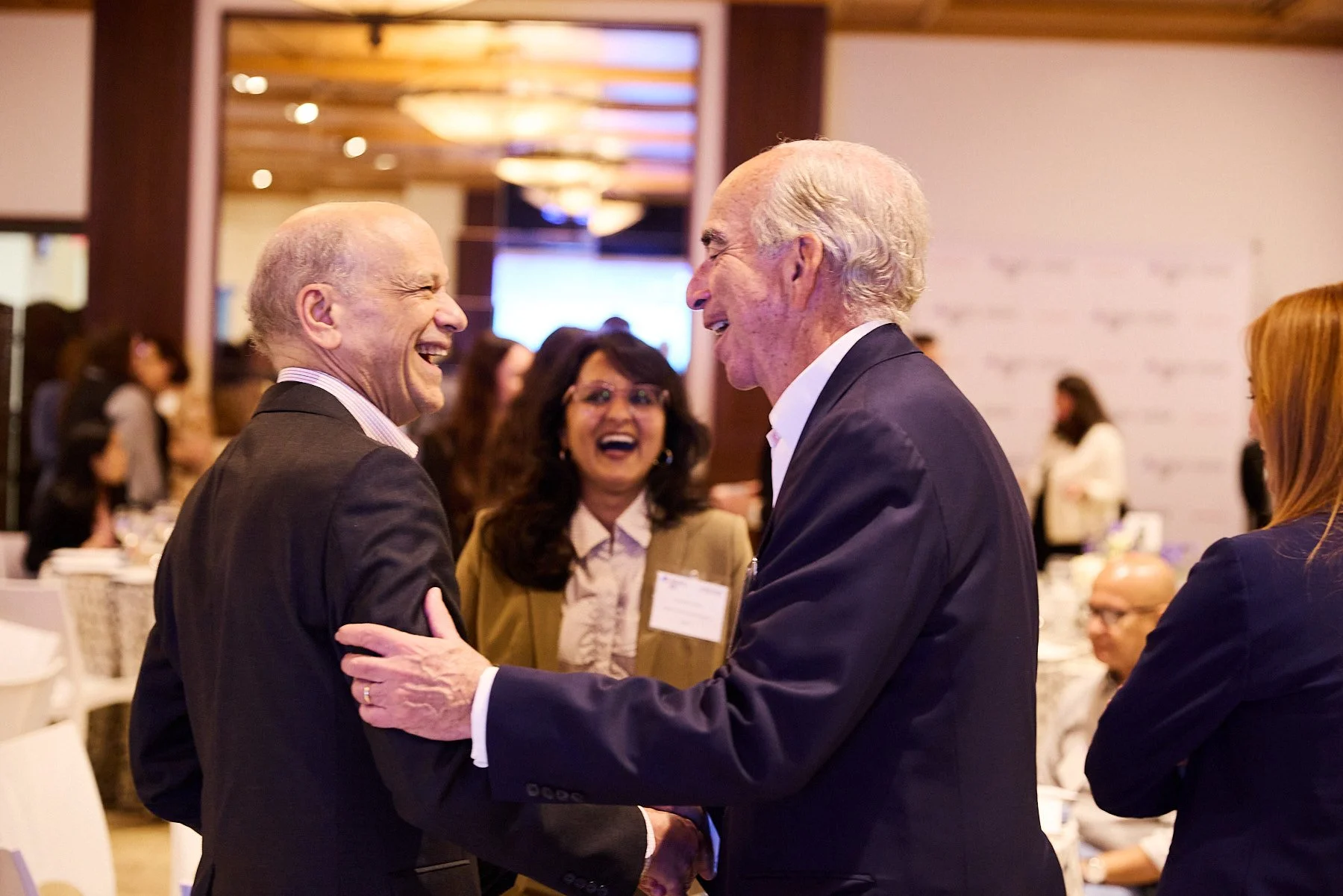 Two older men in suits smiling and shaking hands at a professional event, with a woman with dark hair and glasses smiling in the background.