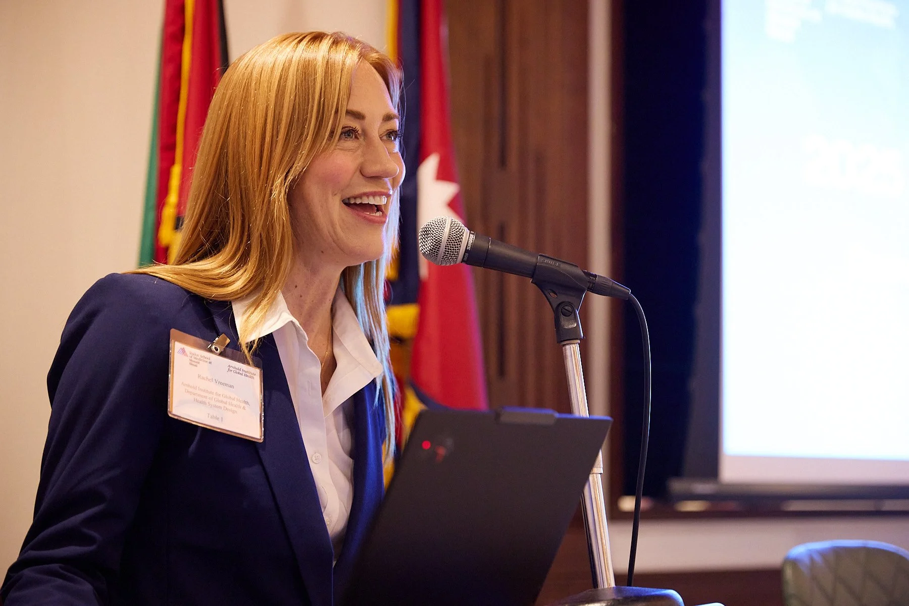 A woman with red hair, wearing a dark blazer and white shirt, speaking into a microphone at a presentation or conference. She has a name badge on her blazer. There are flags in the background and a large screen to her right.
