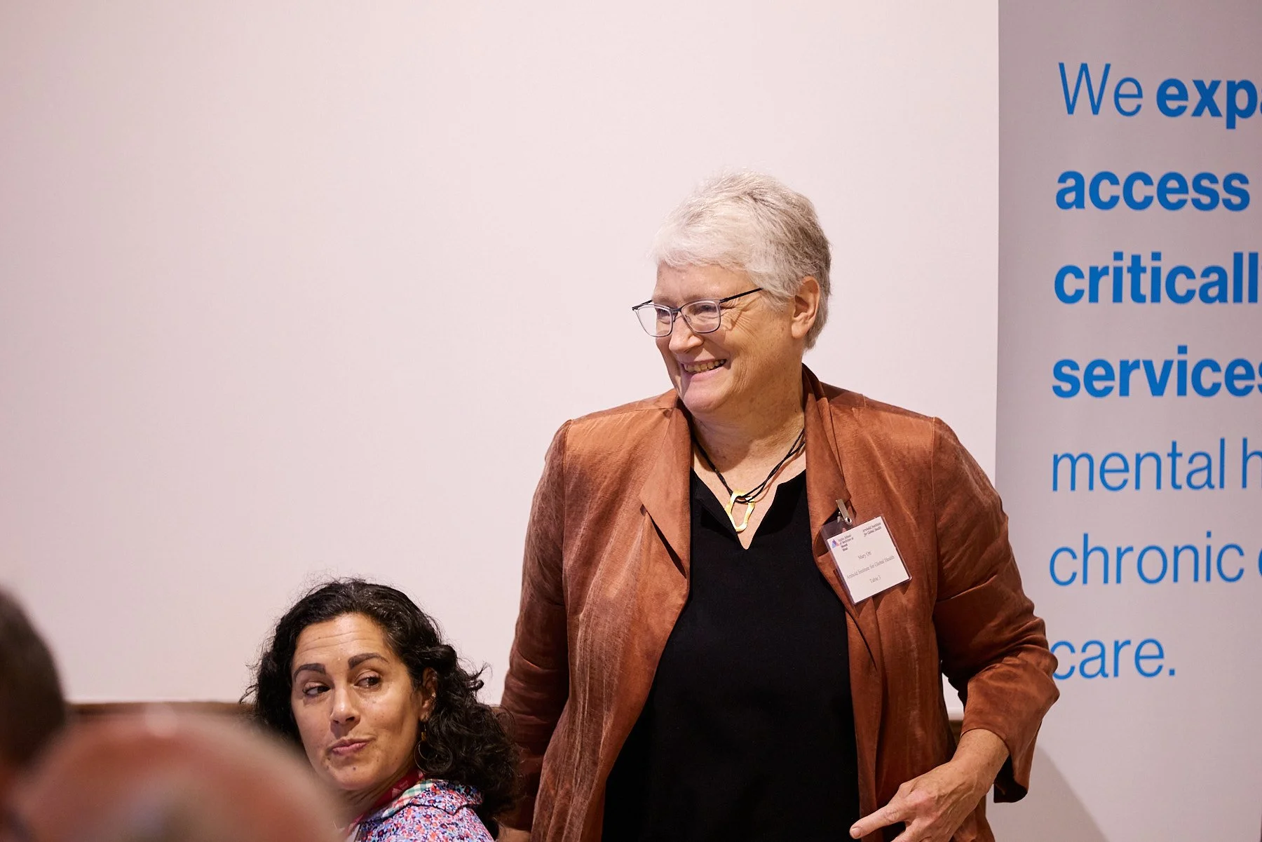 Two women are at a professional event, with one woman smiling and standing, wearing glasses, a brown jacket, and a black top, and the other woman sitting, with dark curly hair, wearing a patterned shirt, looking to the side. In the background, a larg