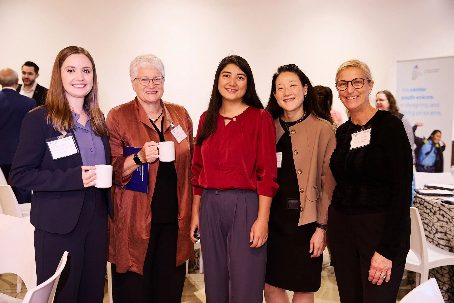 Five women standing together at a professional event, smiling and holding coffee cups, with nametag badges visible.